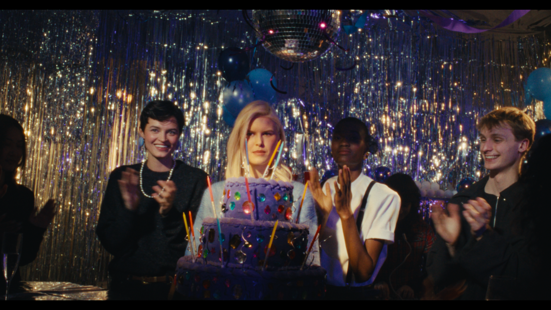 A woman celebrating her birthday at a party with friends, surrounded by gold and blue decorations, with a purple birthday cake topped with candles and colorful decorations in front of her.