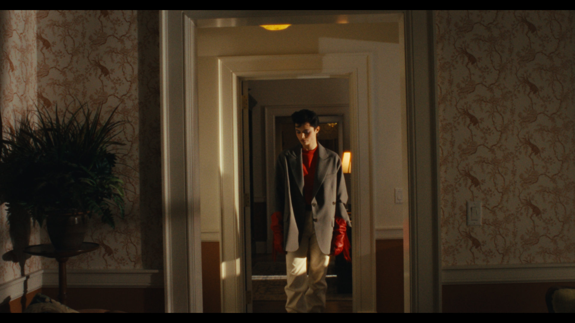 A person with short dark hair wearing a beige blazer, red shirt, beige pants, and red gloves, standing in a dimly lit doorway in a home with floral wallpaper, a potted plant on a side table, and warm lighting in the background.