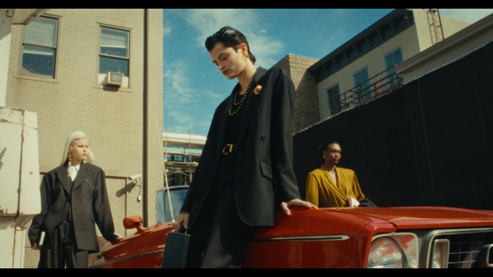Three women stand around a red car in an urban setting on a sunny day, with buildings and a clear sky in the background.