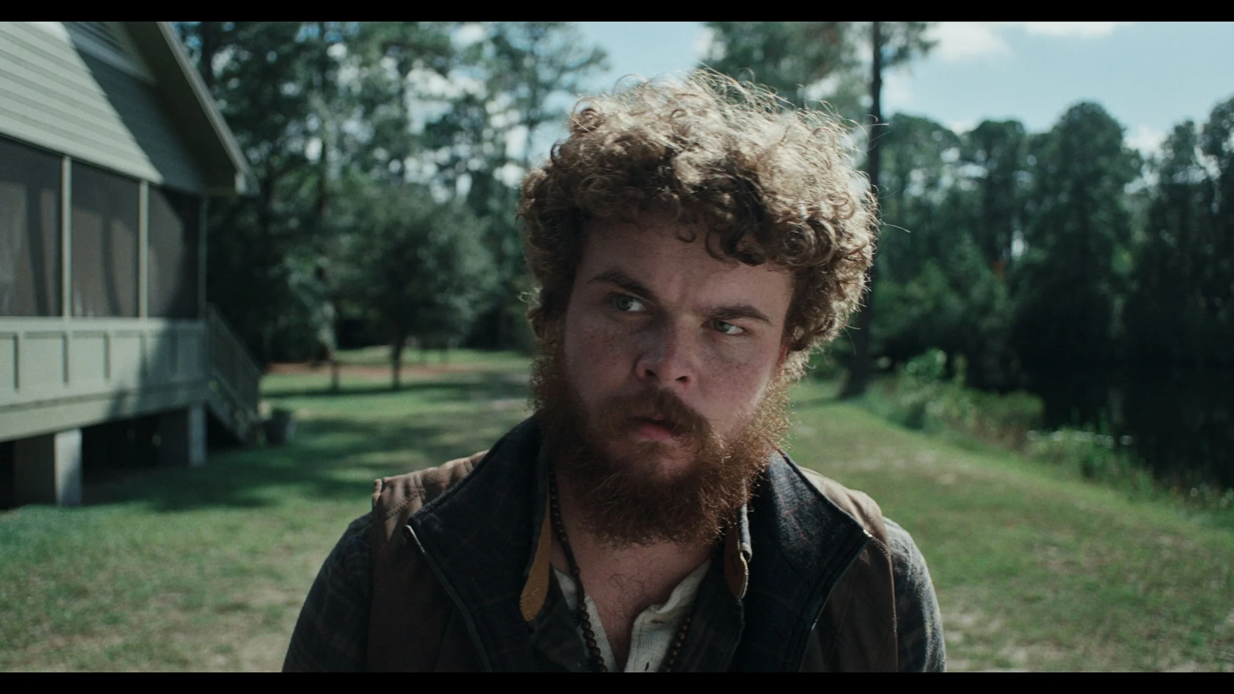 A man with curly hair, a beard, and freckles looks to the side outdoors near a house with trees in the background.