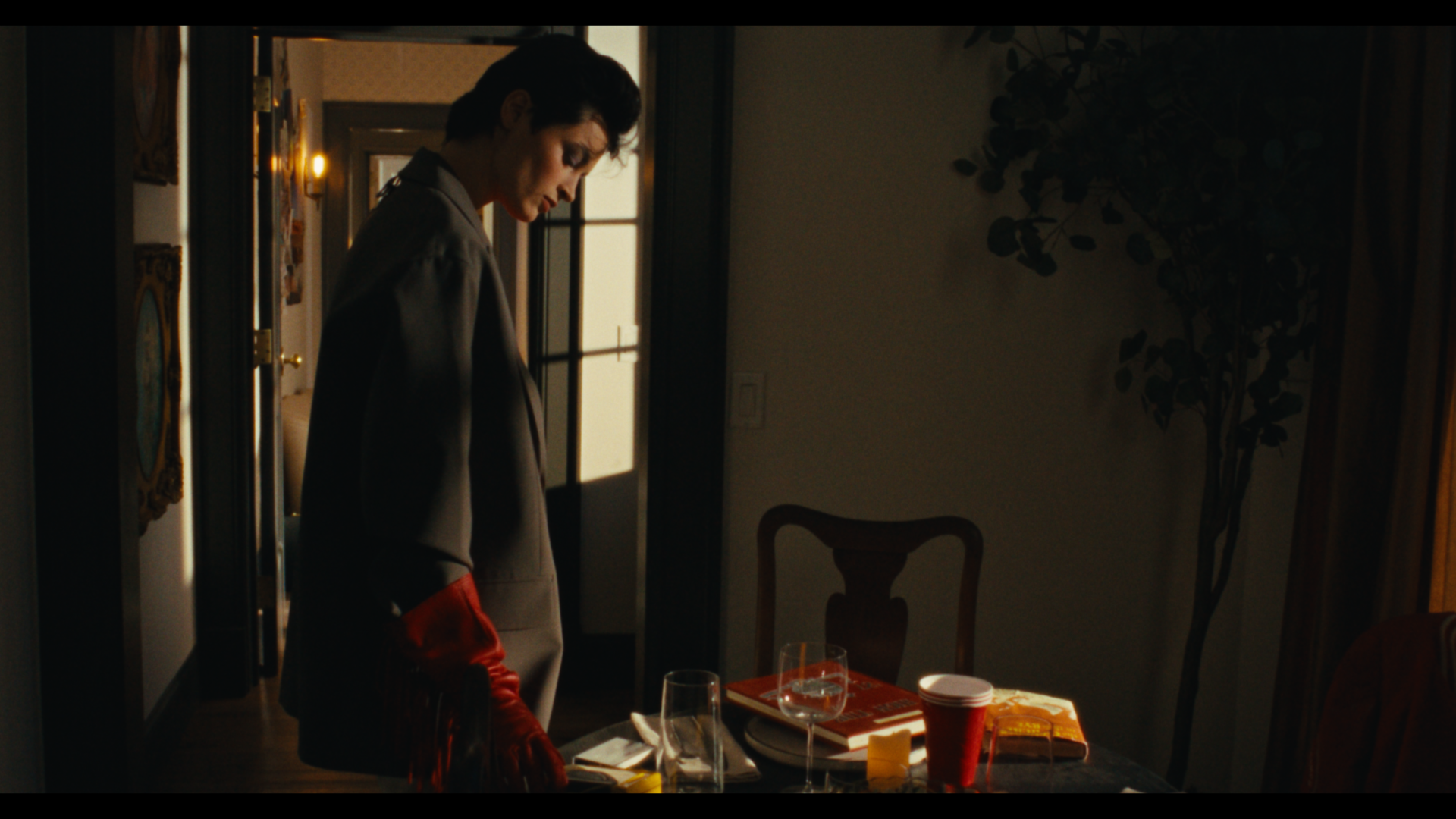 A woman with short dark hair, wearing a gray blazer and red gloves, stands in a dimly lit room near a dining table set with wine glasses, a red cup, books, and snacks, looking downward. The room has a door with sunlight coming through, and a potted p