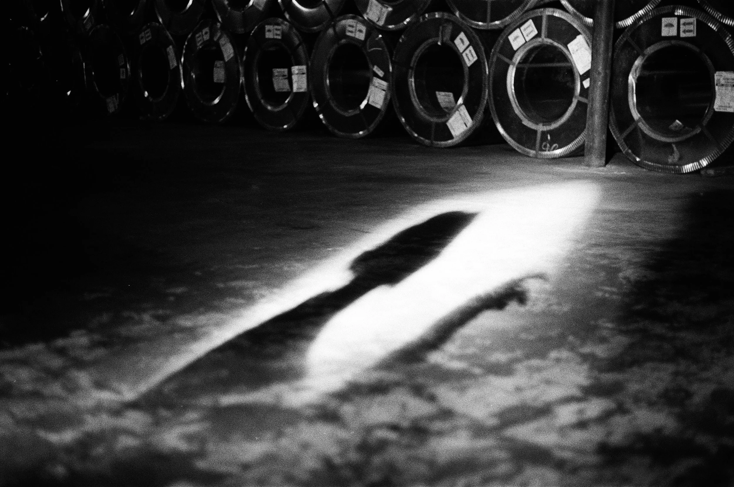A shadow of a hammer cast on the floor in front of a row of large rolls of metal in a dark industrial setting.