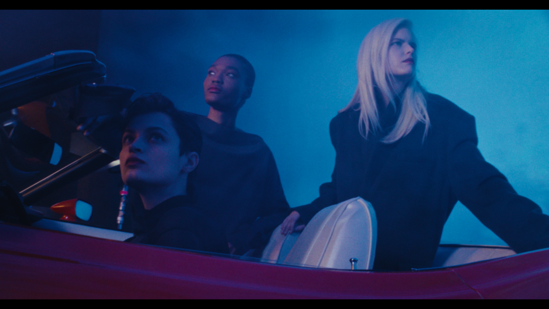 Three women in a vintage red convertible car, with blue and purple lighting creating a moody atmosphere.
