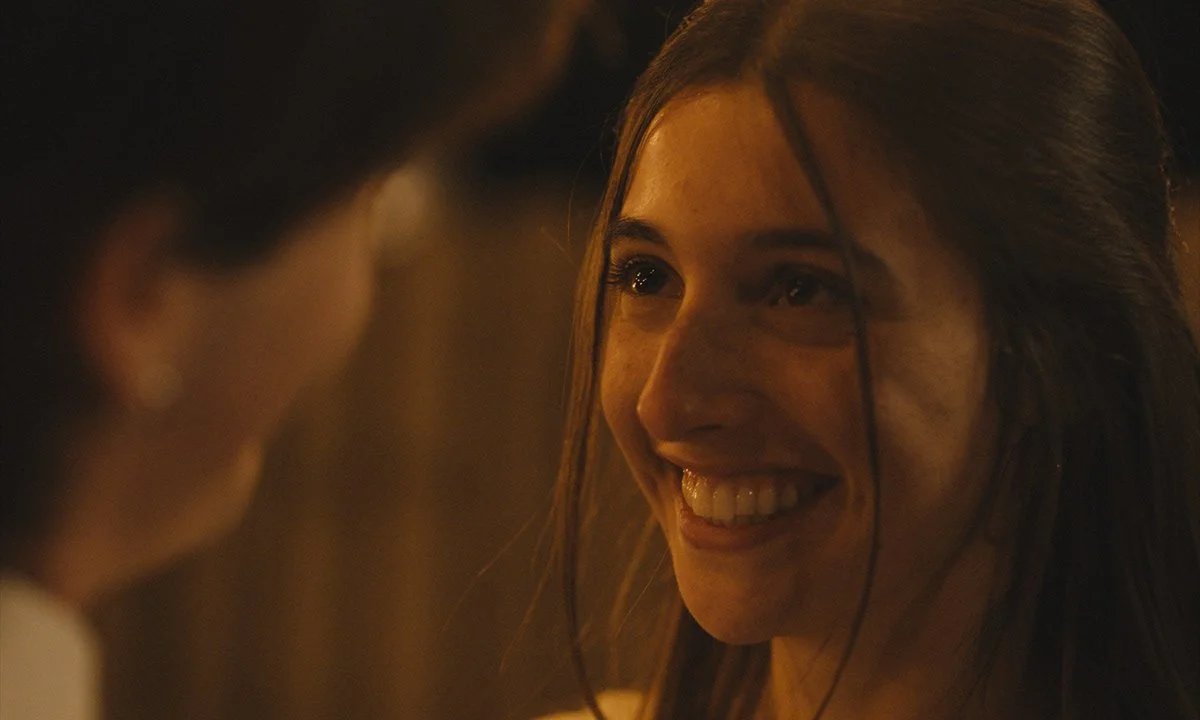 A woman with long brown hair smiling and looking at another person in a dimly lit scene.