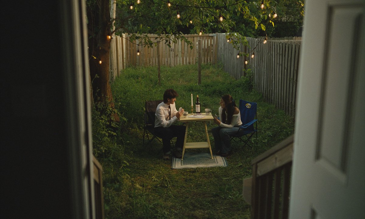A man and woman sitting at a small table in a backyard, having a dinner with wine bottles, candles, and cups, surrounded by string lights and a wooden fence.