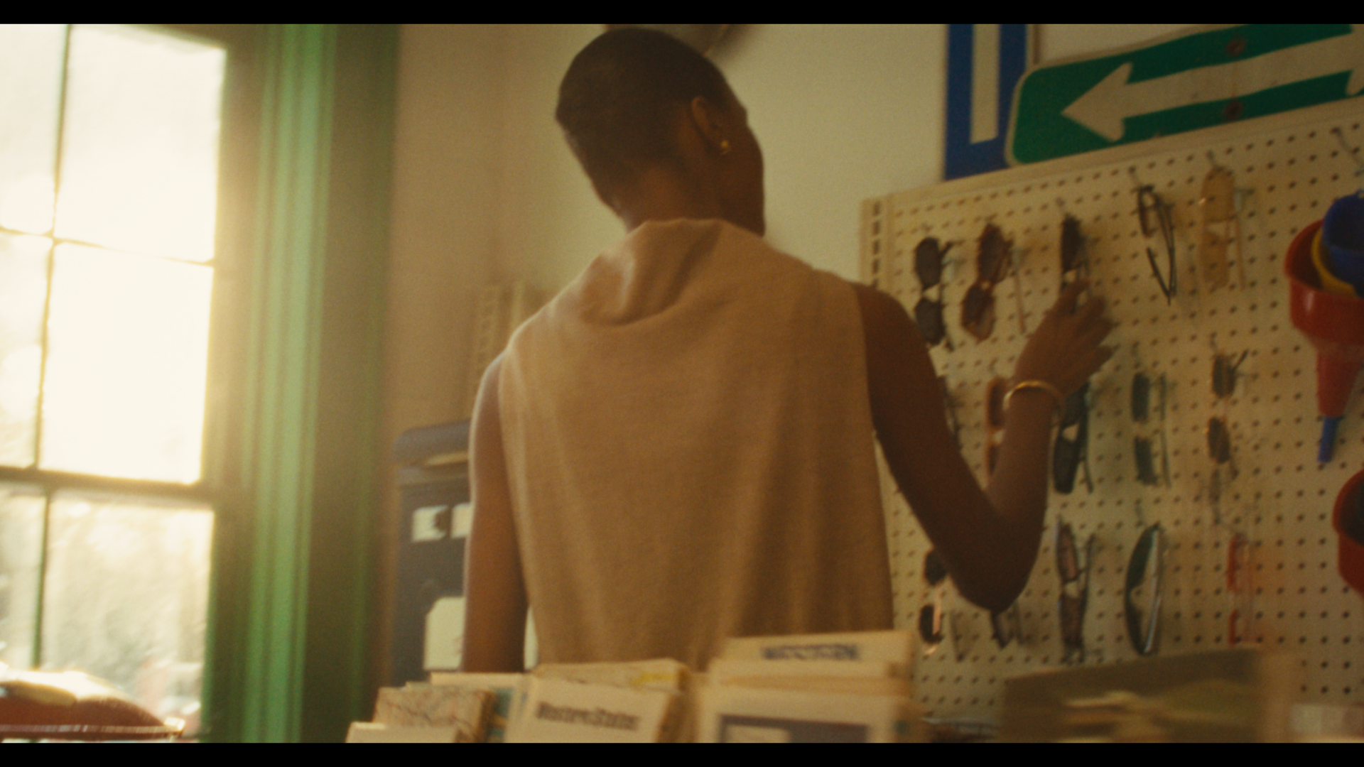 A person shopping for sunglasses in a store, seen from behind, browsing a display of sunglasses on a pegboard. The store is lit with natural sunlight.
