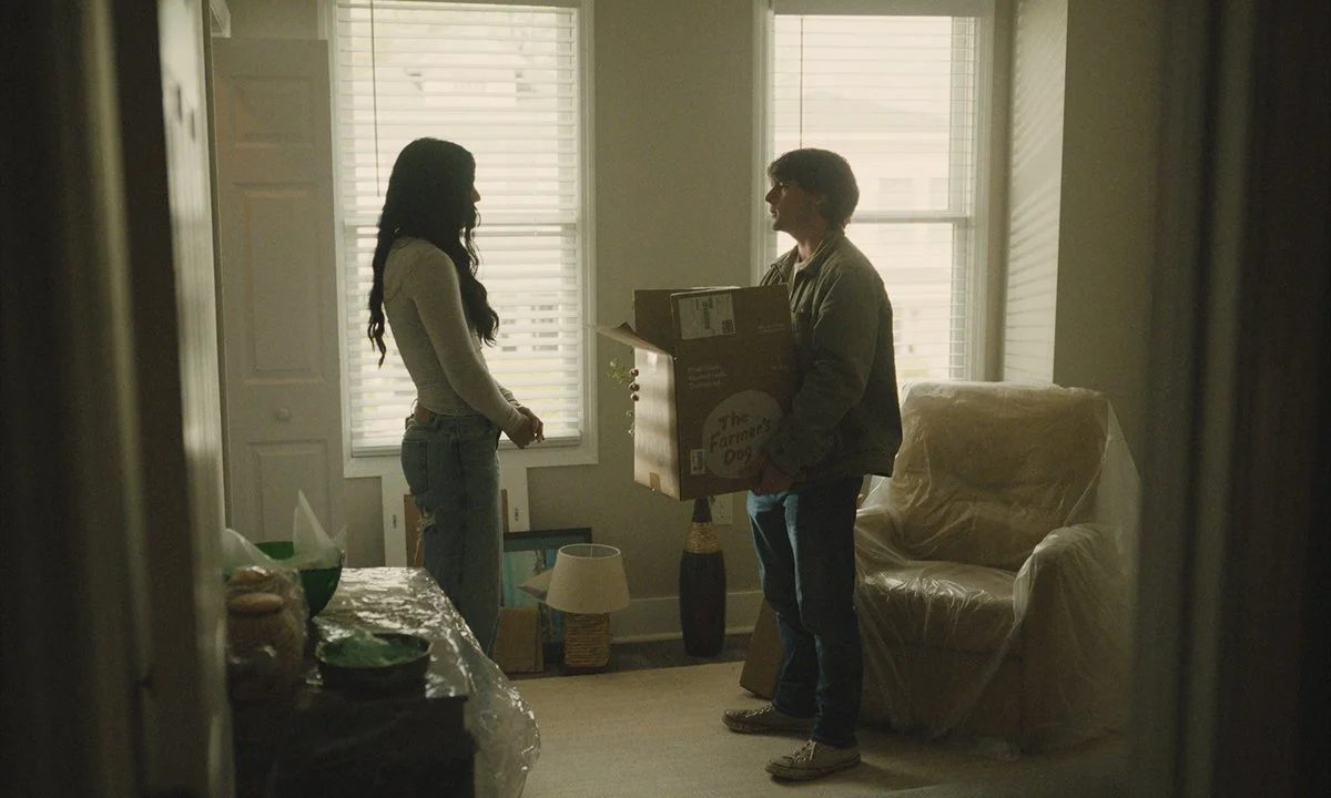 A young man handing a box to a young woman in a living room with windows and a covered armchair.