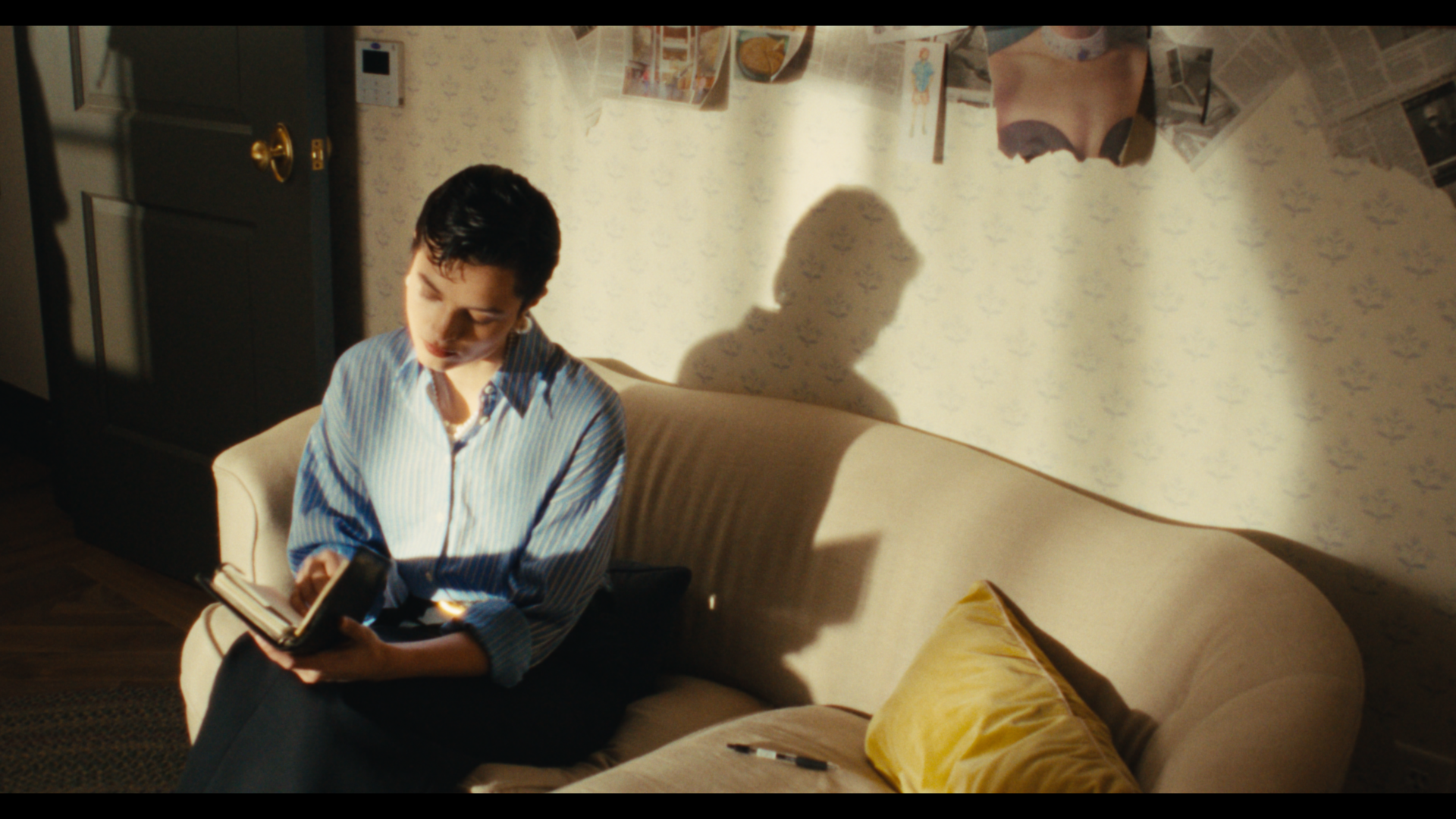 A woman with short dark hair in a striped shirt sitting on a beige couch, looking at a book or tablet, with sunlight casting a shadow of her profile on the wall; a yellow pillow and a black marker are on the couch, and newspaper clippings are pasted 