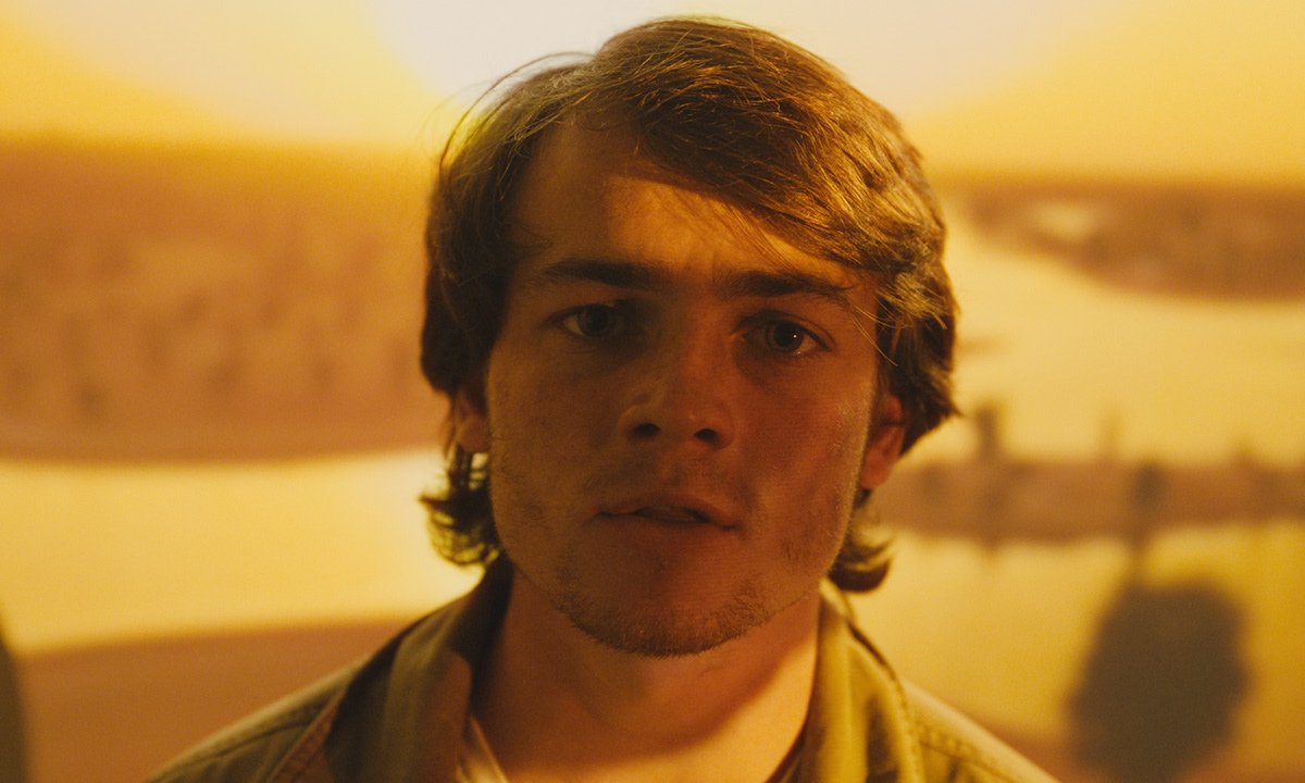 A young man with brown hair and a concerned expression, standing indoors with a yellowish background.