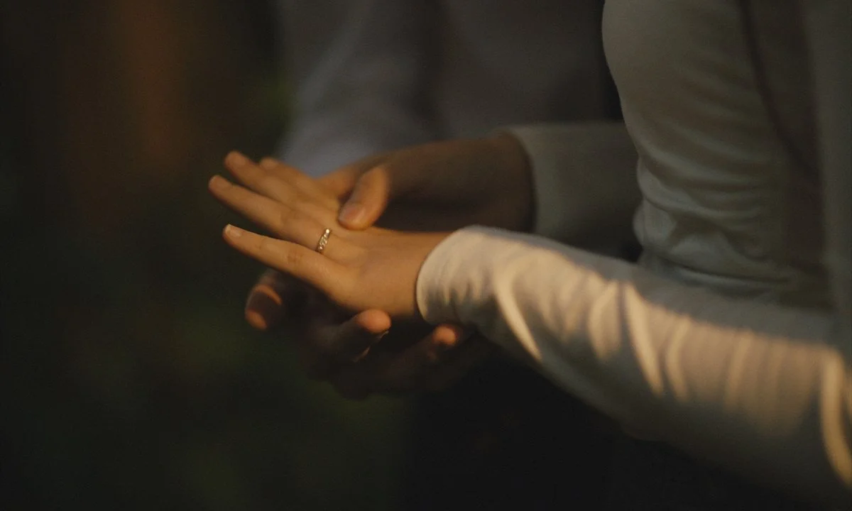 A person wearing a light-colored long sleeve shirt holding another person's hand, showing a wedding ring, in a dark setting.