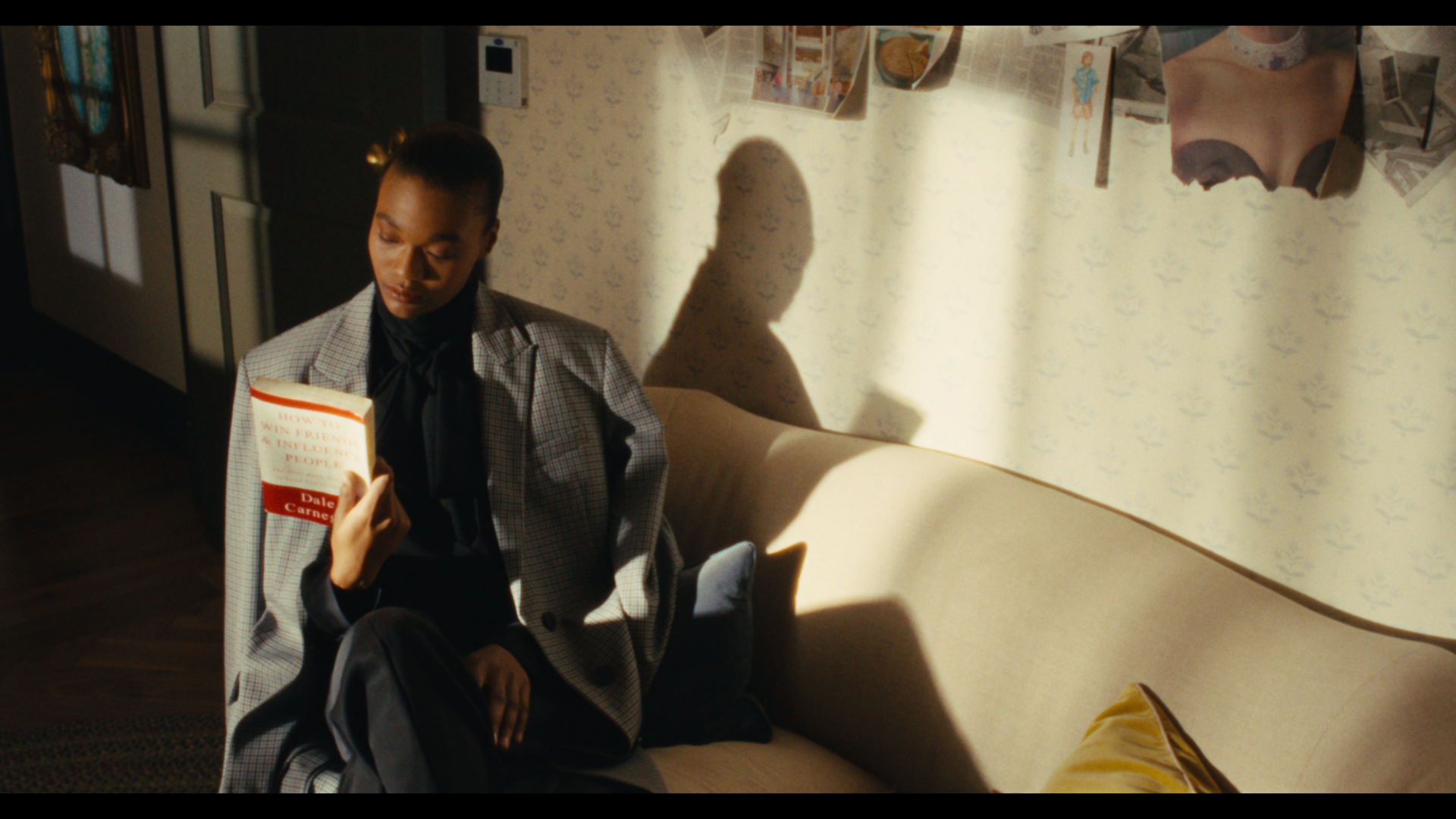 A woman sitting on a beige couch reading a book titled "How to Win Friends & Influence People" in a room with patterned wallpaper, a yellow pillow on the sofa, and a collage of photographs and magazine clippings on the wall, with sunlight casting sha