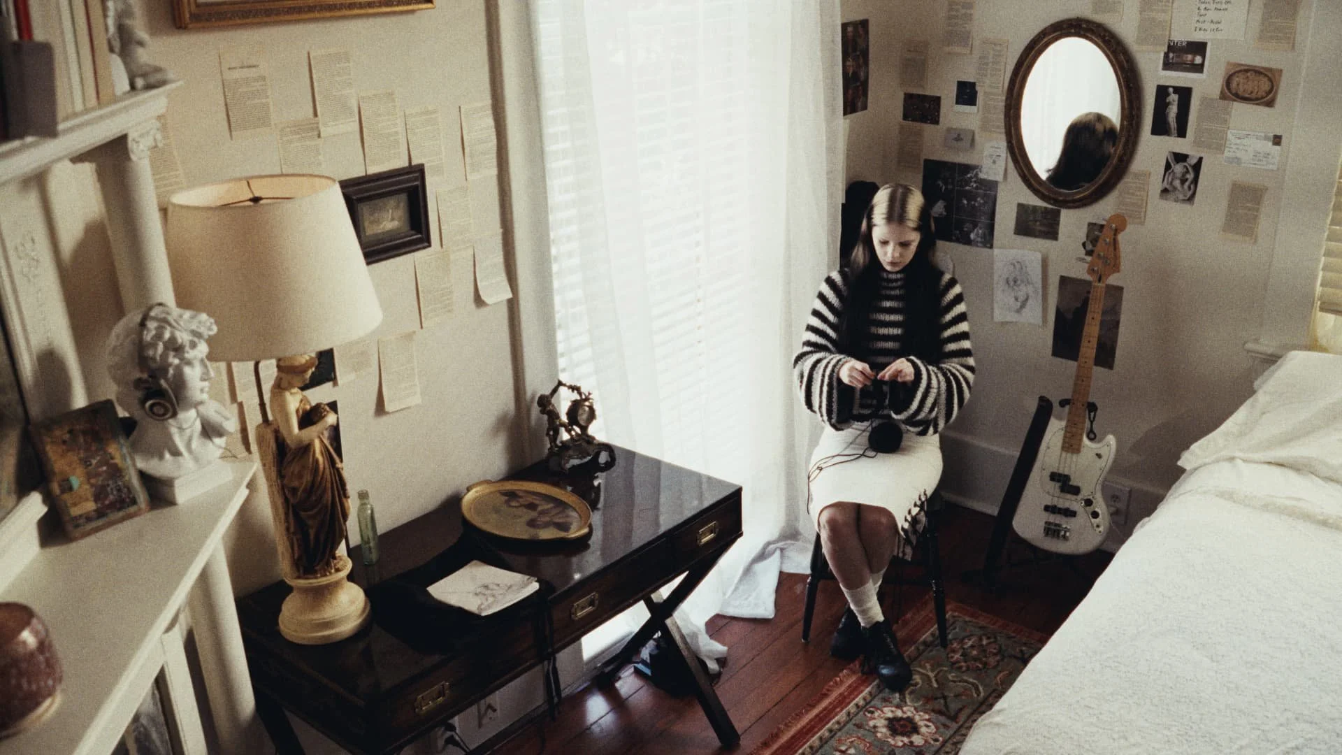 A young woman with long dark hair, wearing a black and white striped sweater and a white skirt, sits on a black chair in a decorated bedroom, listening to music with headphones, holding a music player. The room has a wall with books, papers, and pict