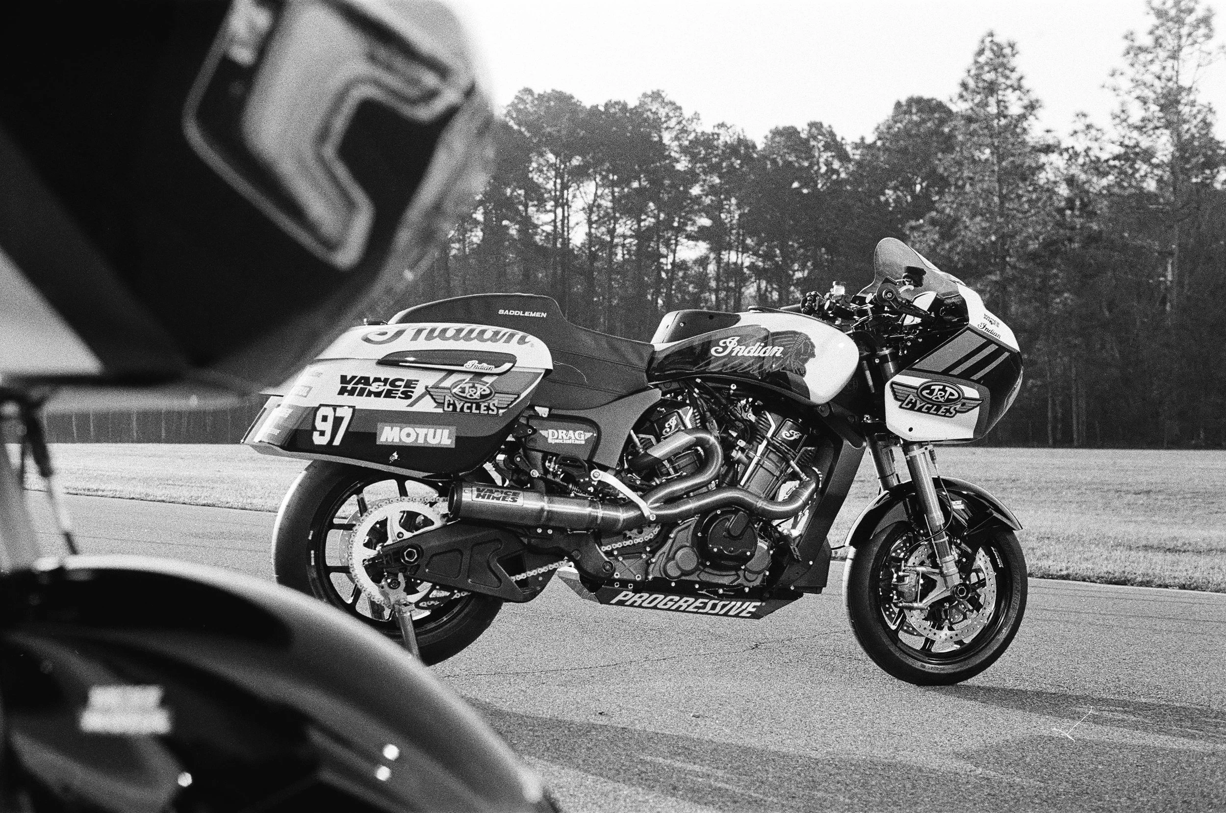 Black and white photo of an Indian racing motorcycle with race number 97 on the side, parked on a track with a wooded background. Part of another motorcycle's helmet or body is visible in the foreground. Behind the scenes photo by Mathias White