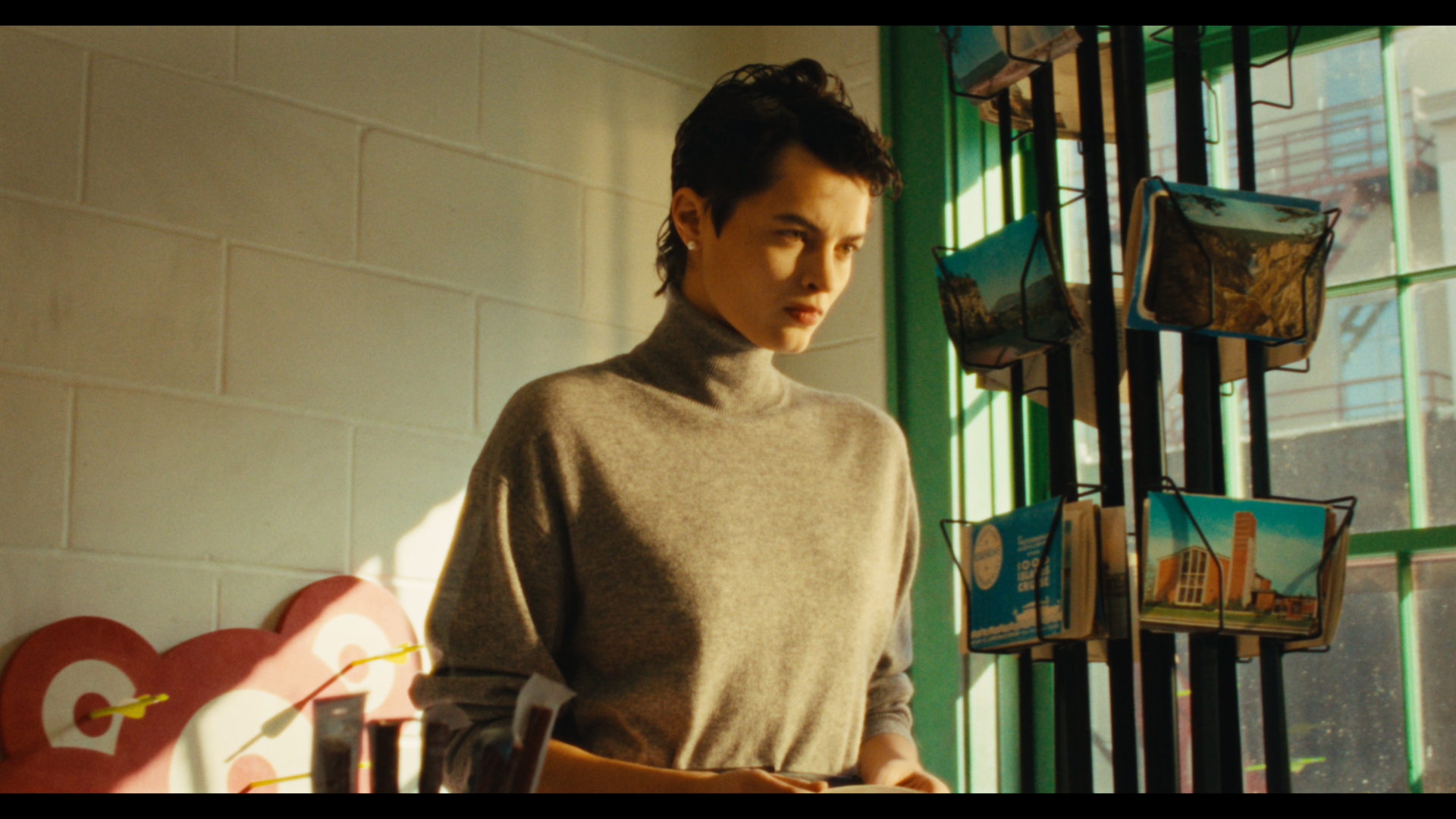 A woman with short dark hair, wearing a gray turtleneck sweater, looking at postcards displayed near a window with sunlight streaming in.