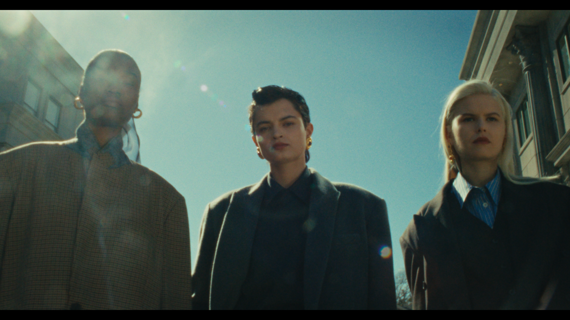 Three women standing under a clear blue sky in an urban setting with buildings behind them. They are dressed in professional attire and looking down at the camera.