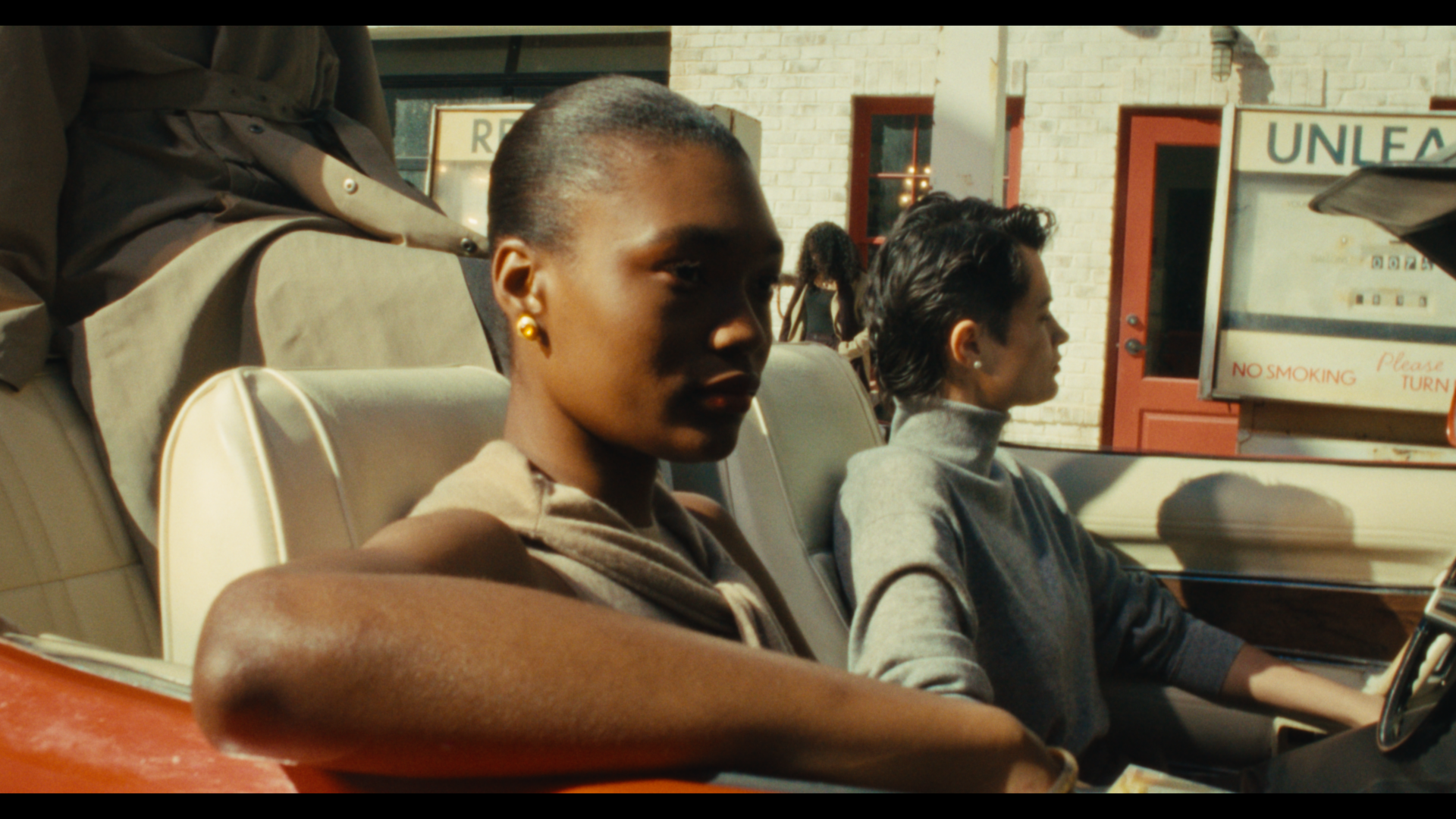 Two women sitting in a vintage convertible car. The woman in the foreground has dark skin, short hair, and is wearing gold earrings and a sleeveless top. The woman in the background has lighter skin, dark short hair, and is wearing a gray turtleneck 