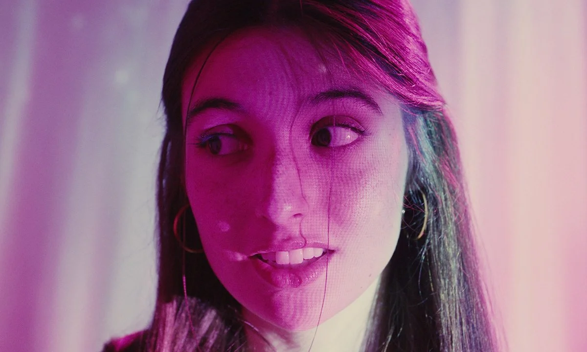 A young woman with long dark hair and hoop earrings, illuminated by pink and purple lighting, looking to one side with a slight smile.