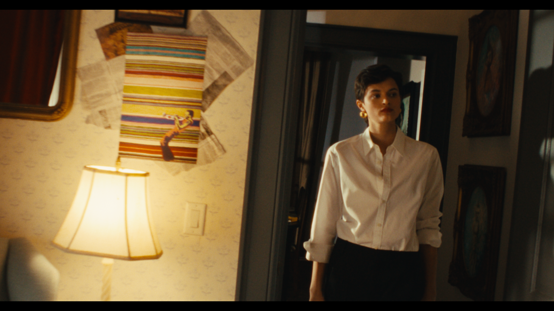 A woman with short dark hair, wearing a white button-up shirt and earrings, standing in a dimly lit room, looking to her right.