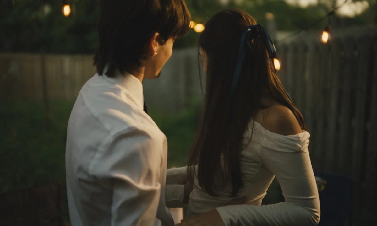 A young man and woman stand close together outdoors during sunset or twilight, with string lights in the background, sharing an intimate moment.