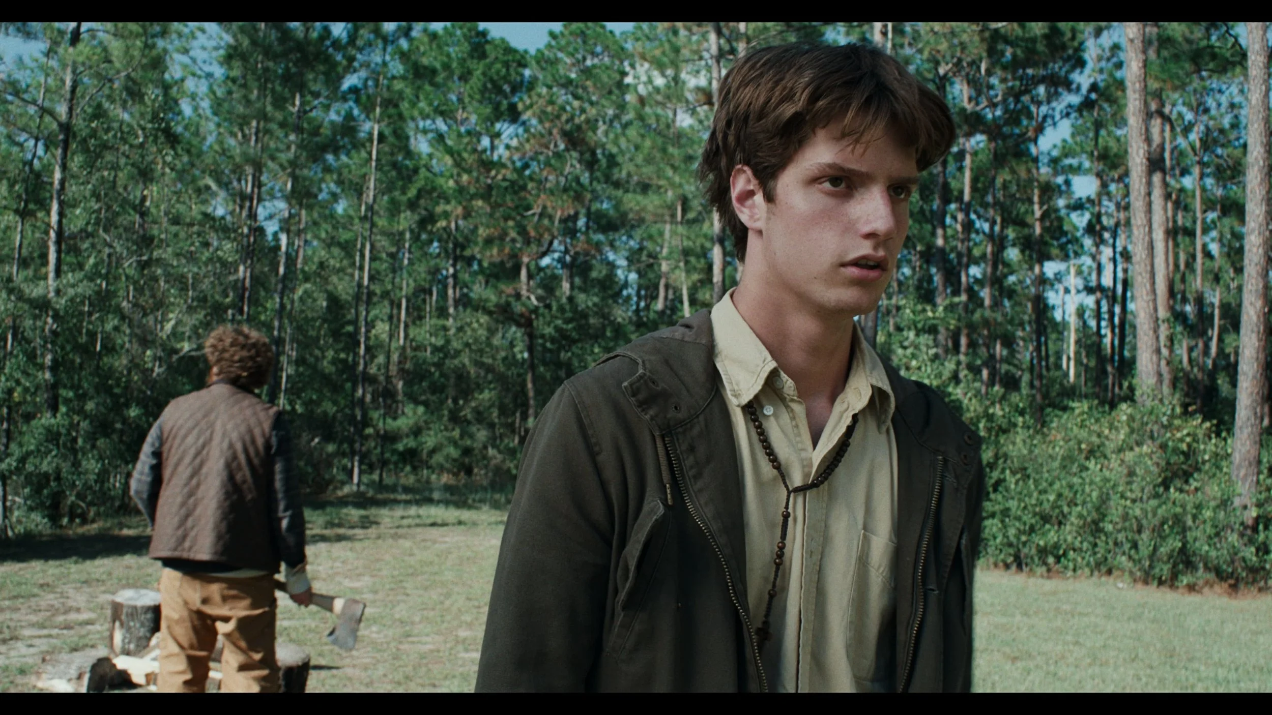 Young man with brown hair wearing a tan shirt, dark jacket, and a beaded necklace standing in a wooded outdoor area, with another person chopping wood in the background.