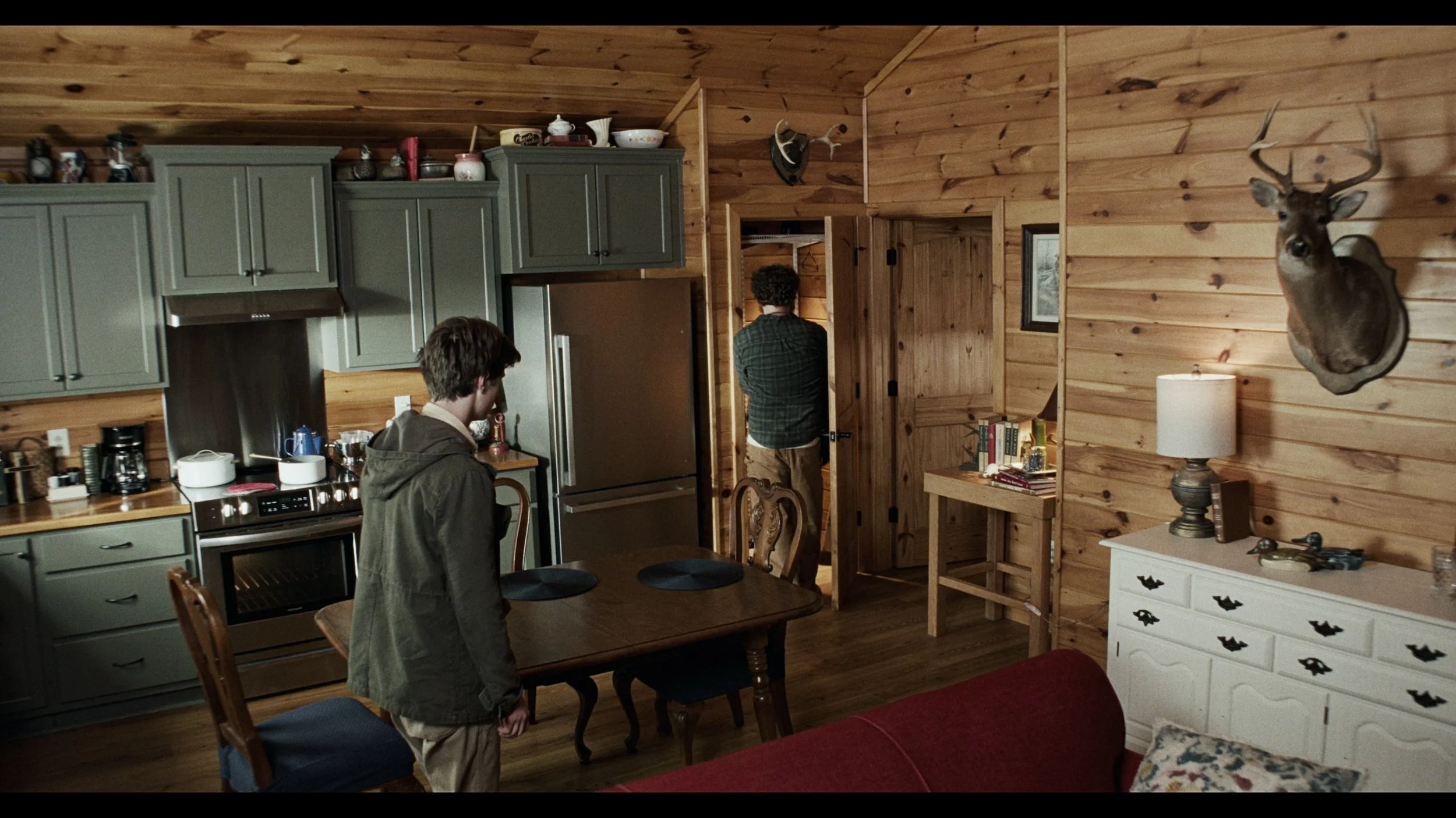 A scene inside a wooden cabin with a kitchen and dining area. Two men are present, one sitting at the dining table and the other standing near a door. The decor includes a mounted deer head, a map on the wall, and various household items and books on