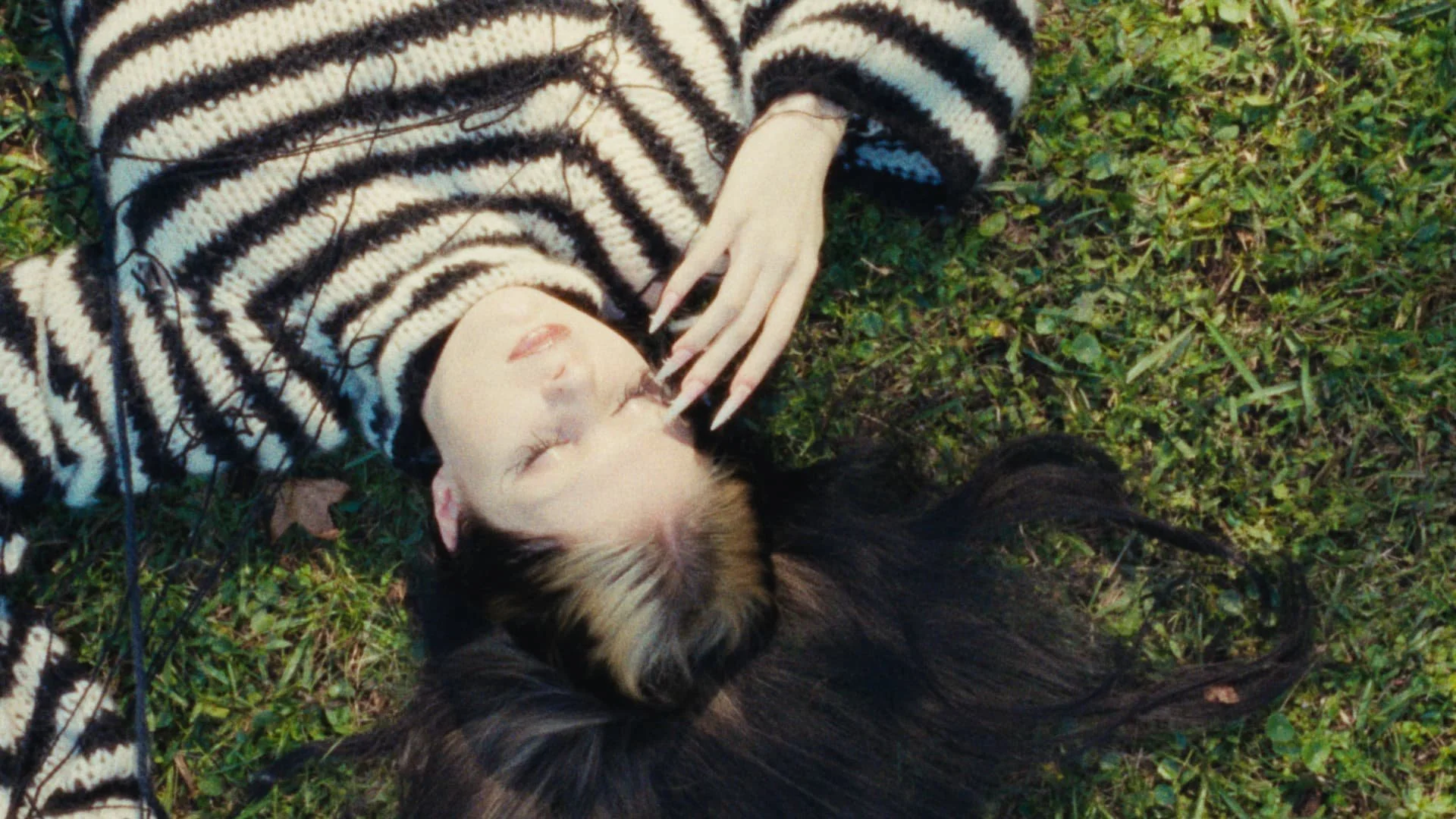 A young woman with long dark hair lying on grass with eyes closed, wearing a black and white striped sweater.