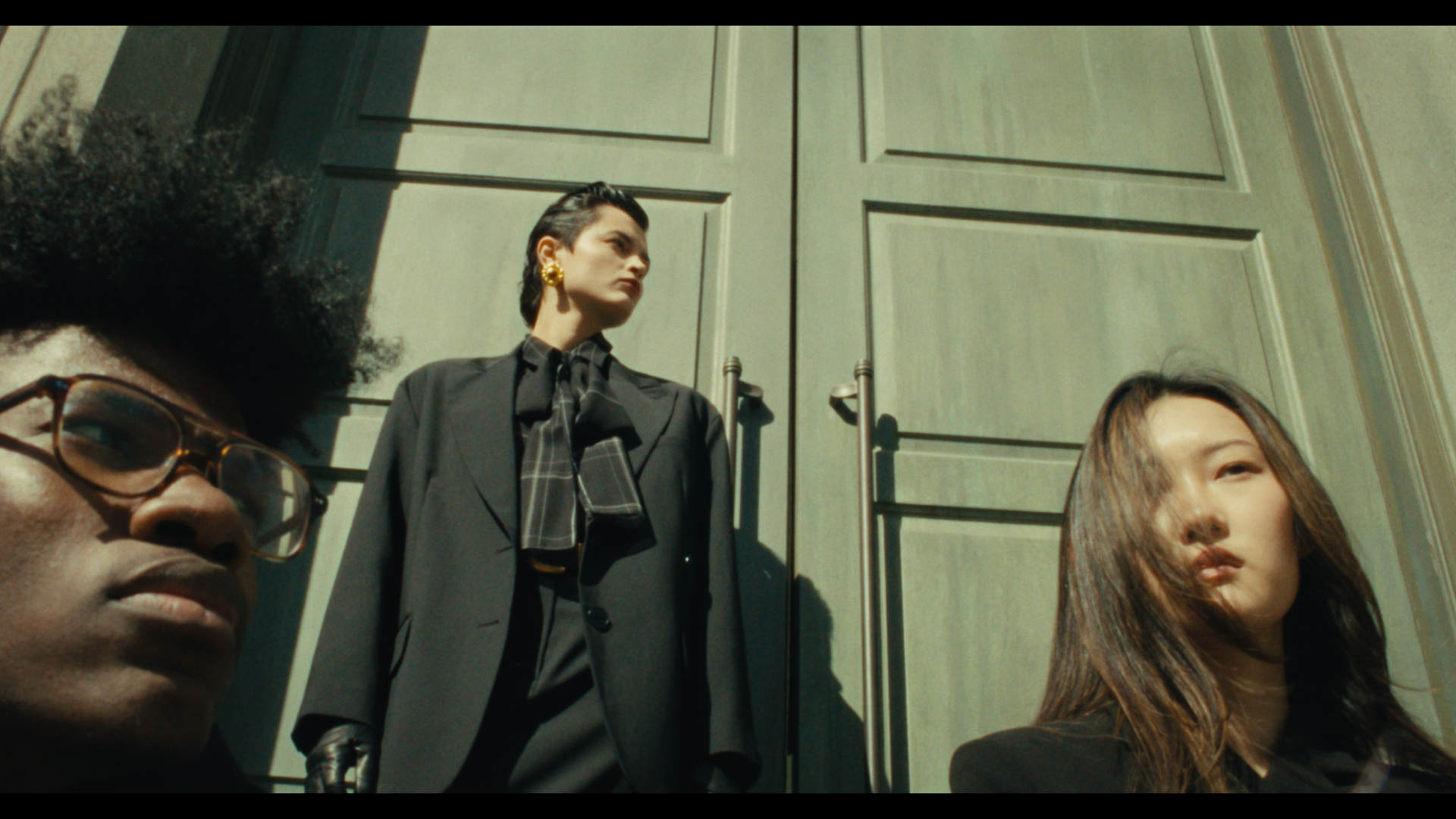 Three women in black formal attire with a green building background