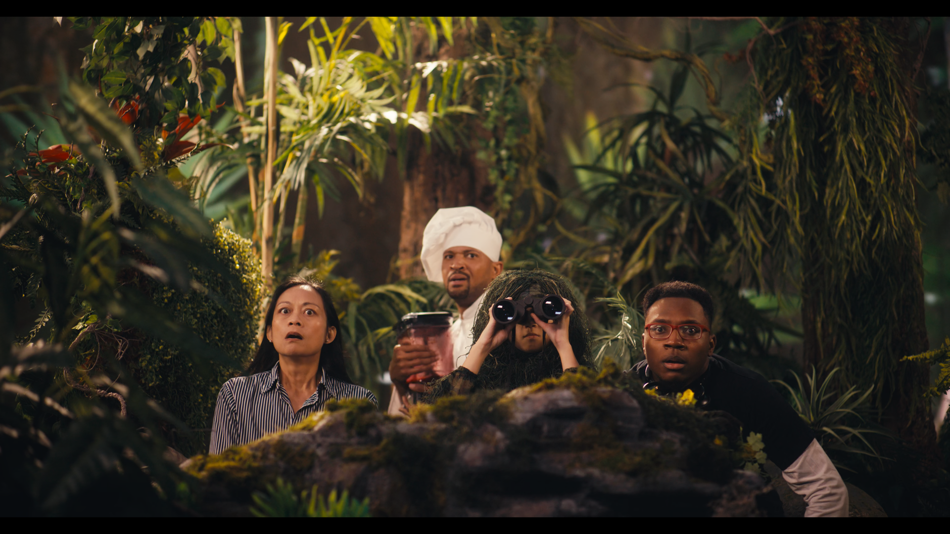 A group of five people with surprised and curious expressions, in a dense jungle setting, looking at something outside the frame. Two are holding binoculars, one is holding a cell phone, and a person wearing a chef's hat is holding a cup. Best Buy ad