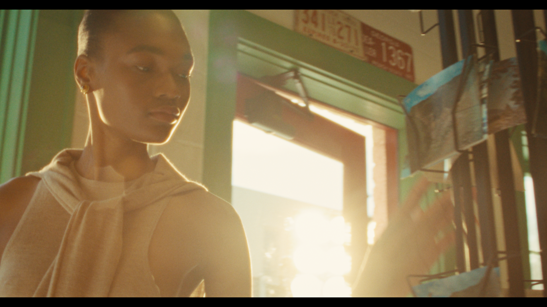 A woman with short hair and hoop earrings stands inside a store, looking at merchandise displayed on a rack, with sunlight streaming through an open door in the background.