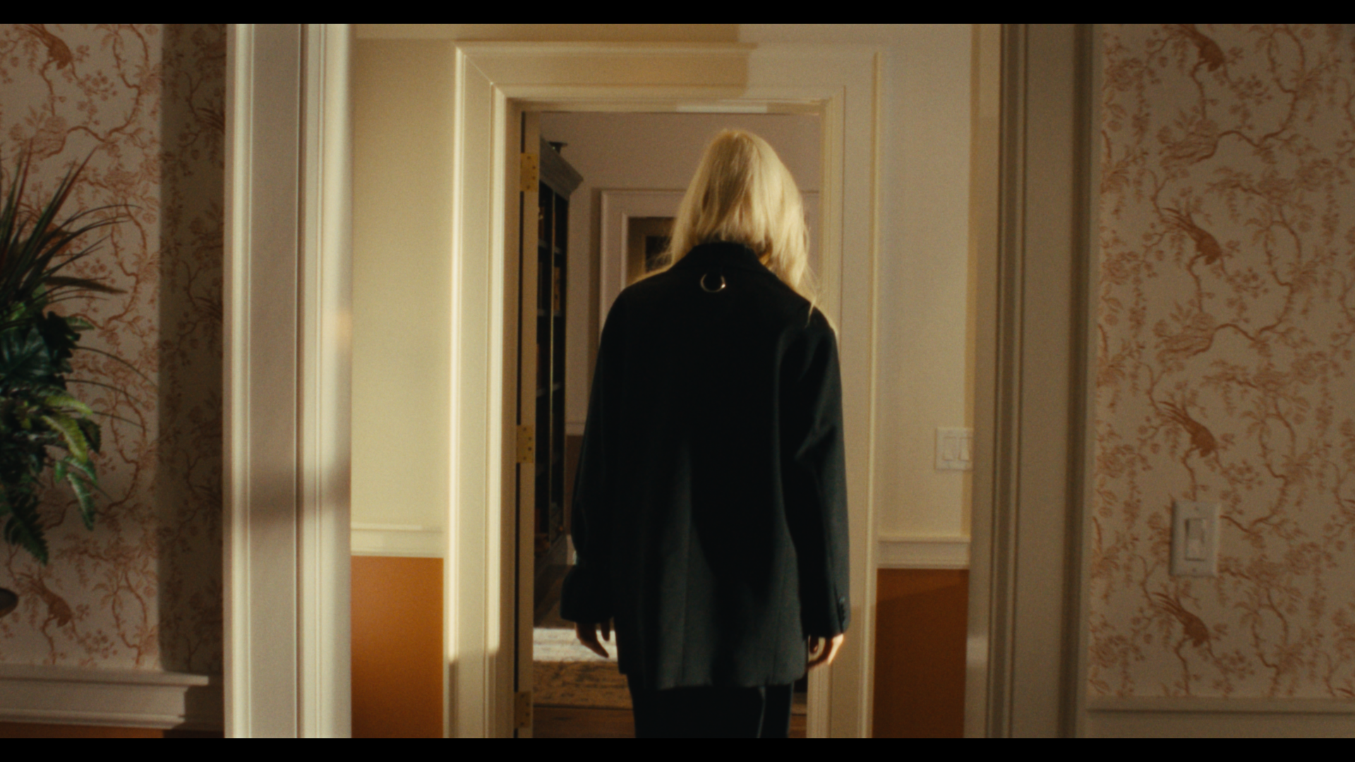 A woman with blonde hair wearing a black blazer walking into a room with yellow and beige walls and floral wallpaper.