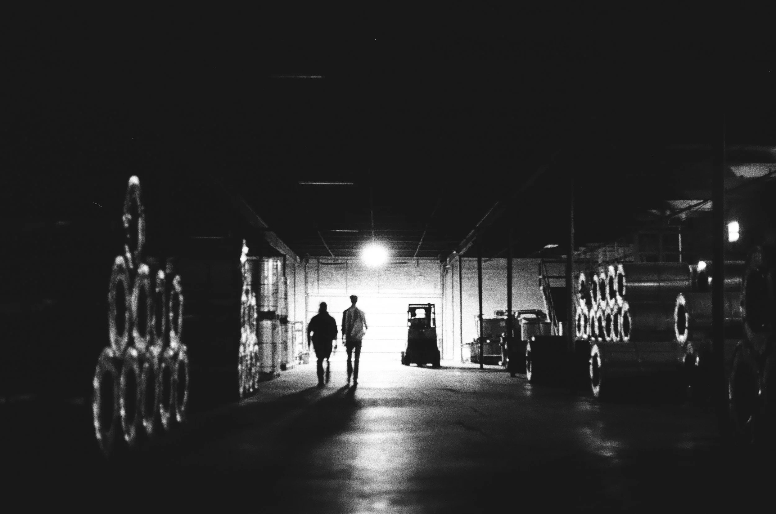 Two people walking side by side in a warehouse or industrial storage area, silhouetted against a bright light at the end of the aisle, with shelves of industrial rolls or drums on both sides.