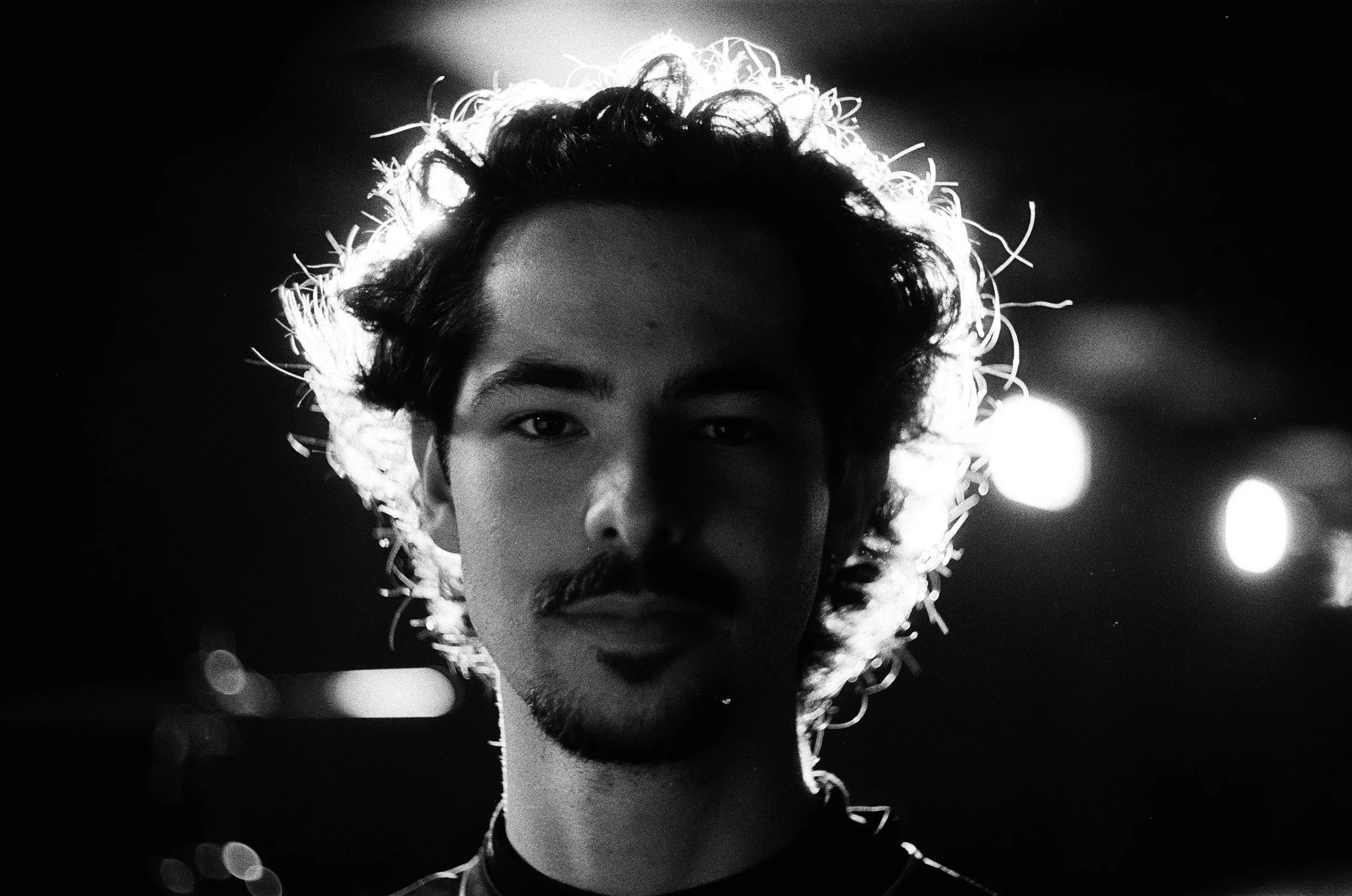 Black and white photo of a man with curly hair and a mustache, backlit to create a halo effect around his head, with blurred lights in the background.
