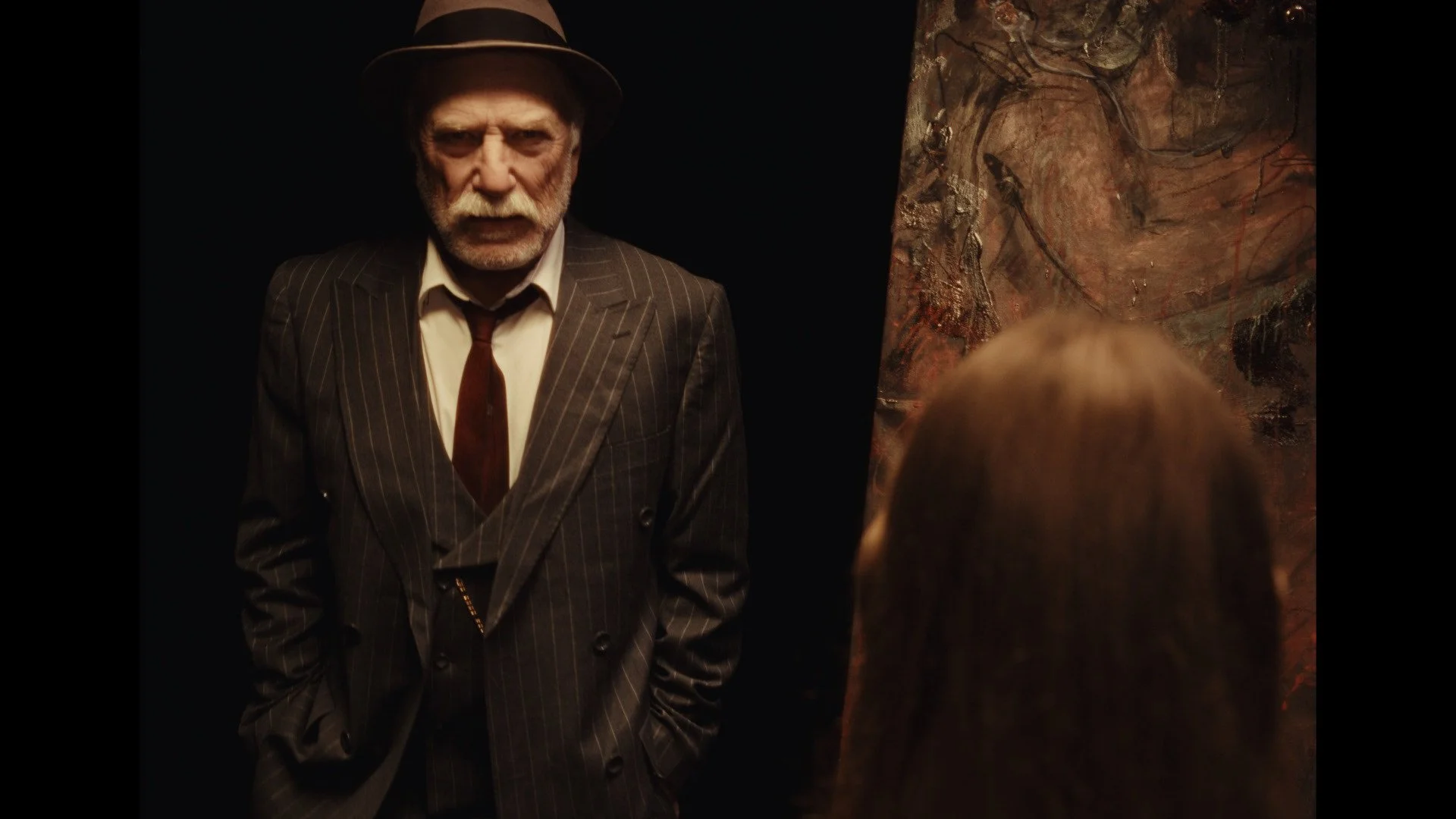 An elderly man with white hair, a beard, and a mustache, wearing a fedora hat, a pinstripe suit, a white shirt, and a brown tie, stands with hands in pockets, looking at a woman with long hair and a painting or textured artwork on the wall behind her