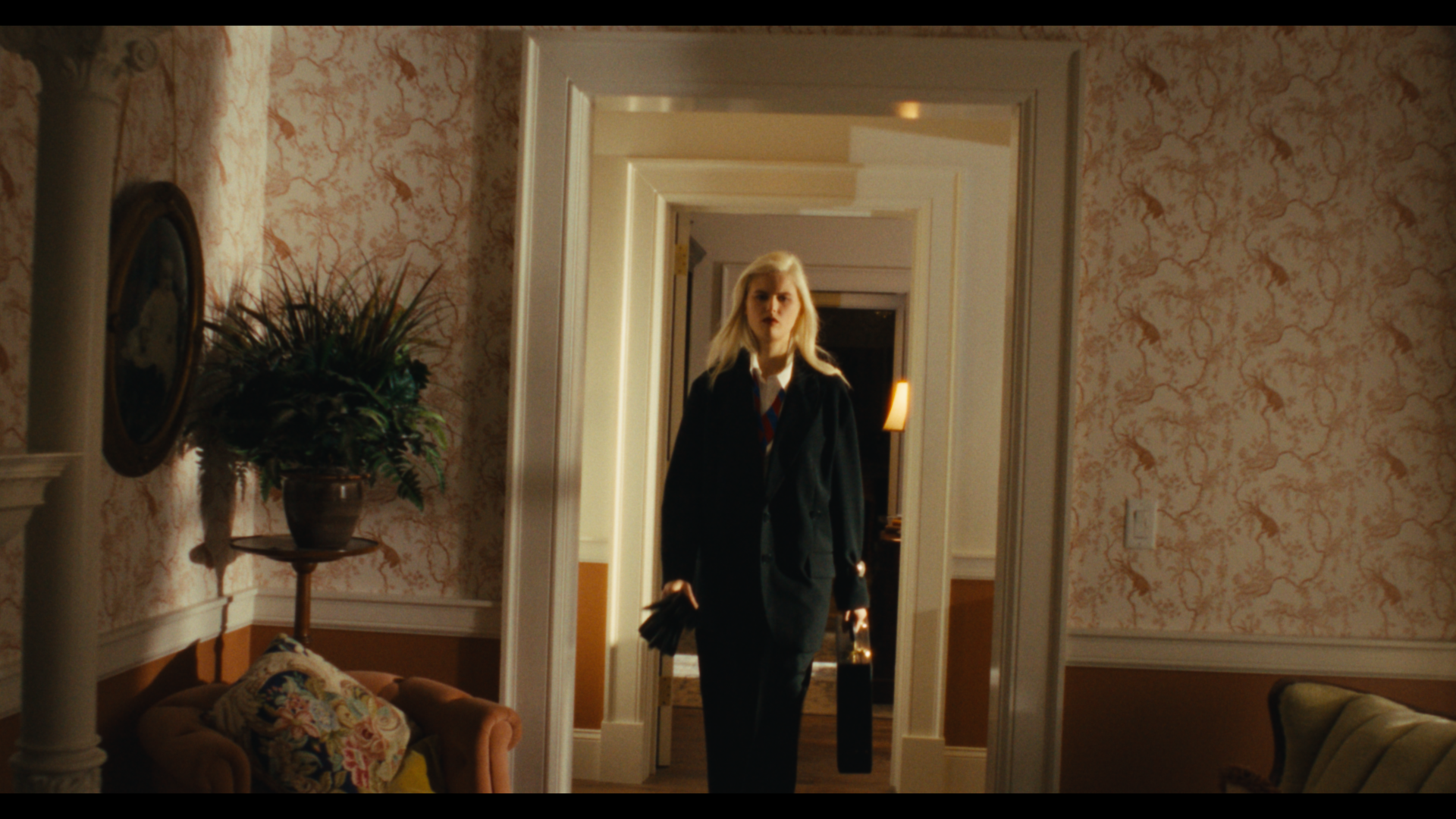 A woman with blonde hair walking through a doorway in a vintage-style room, holding a purse and a small bag, with floral wallpaper and furniture.