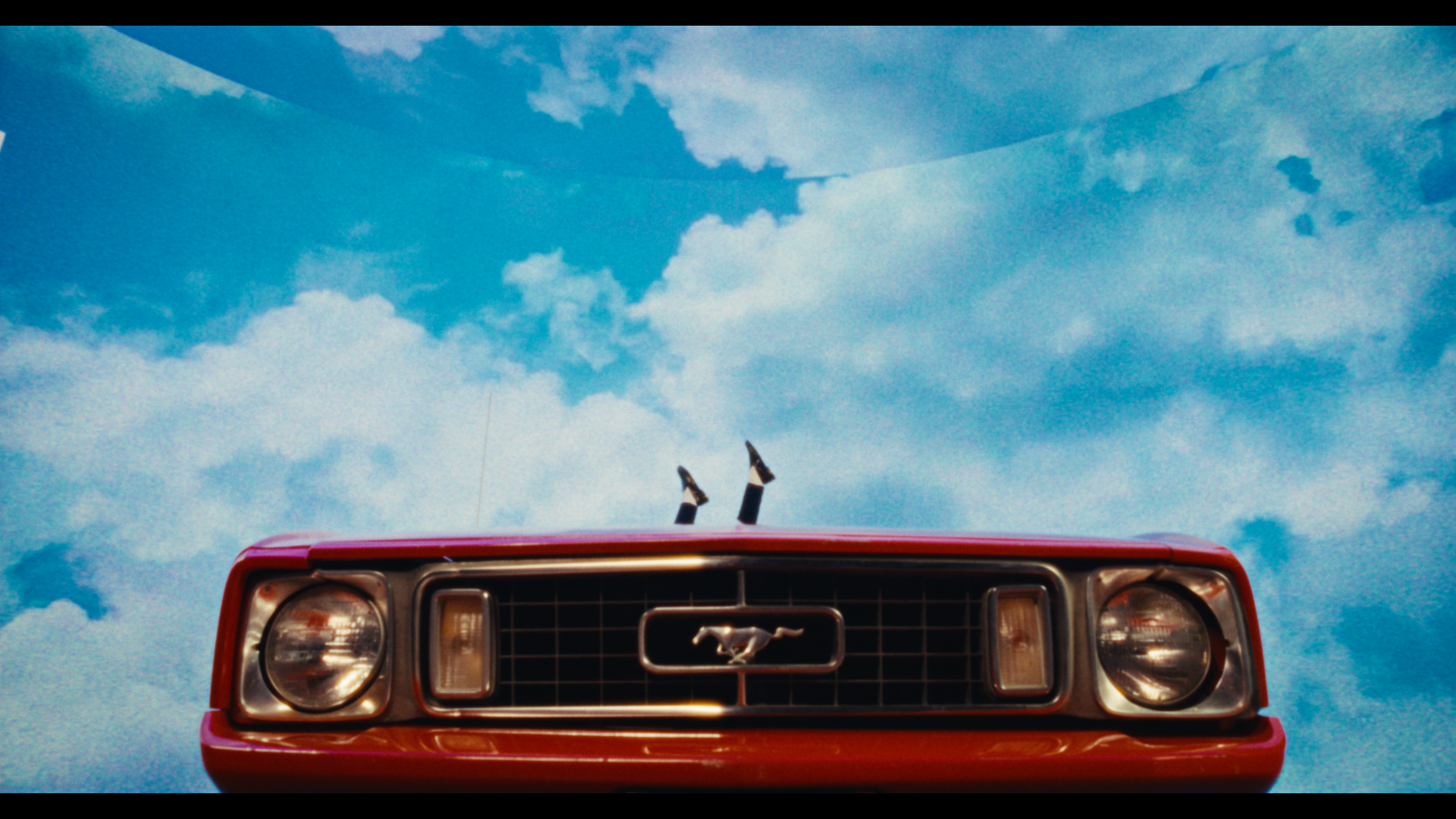 Front view of a red classic Ford Mustang car with a Mustang emblem on the grille, set against a blue sky with fluffy clouds.