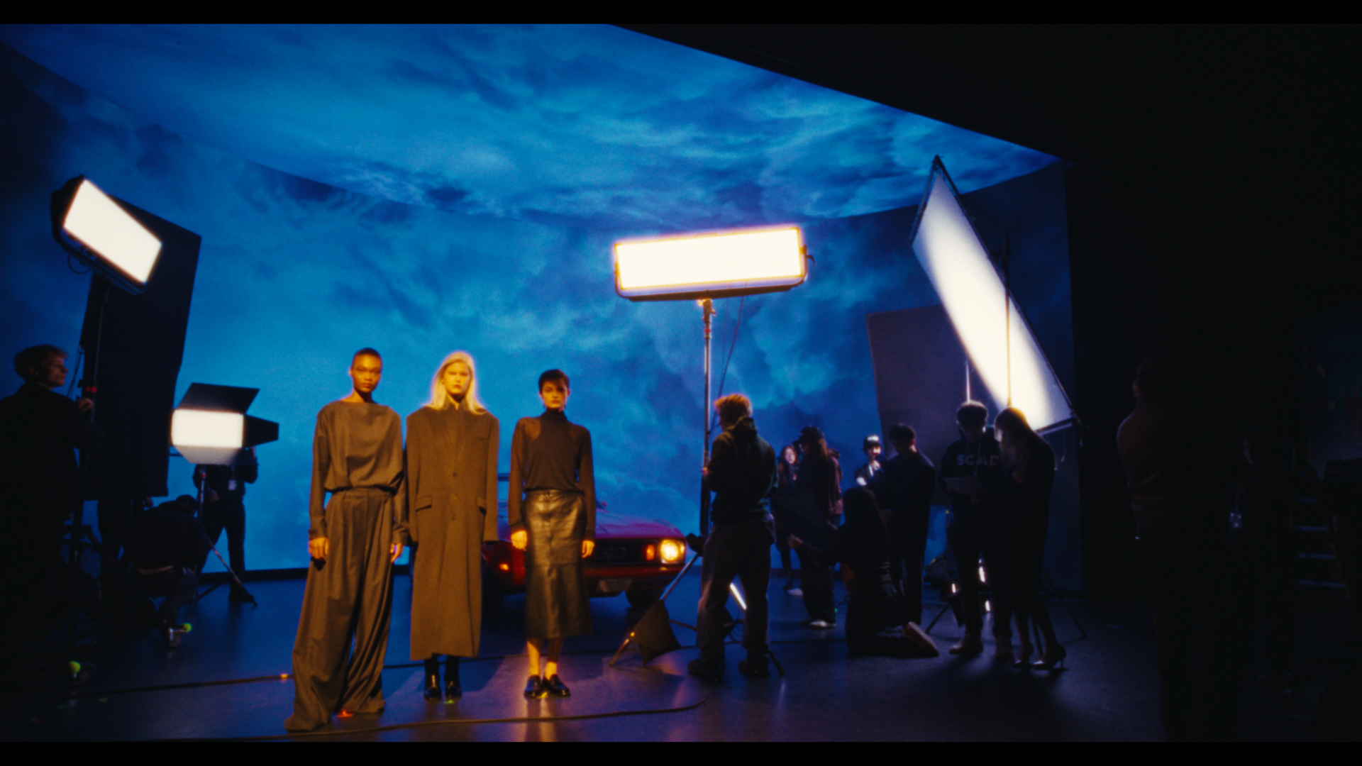 A behind-the-scenes look at a fashion photoshoot in a studio with a large blue sky backdrop. Three models dressed in dark clothing stand in front of a red car, with crew members setting up lighting and equipment around them.