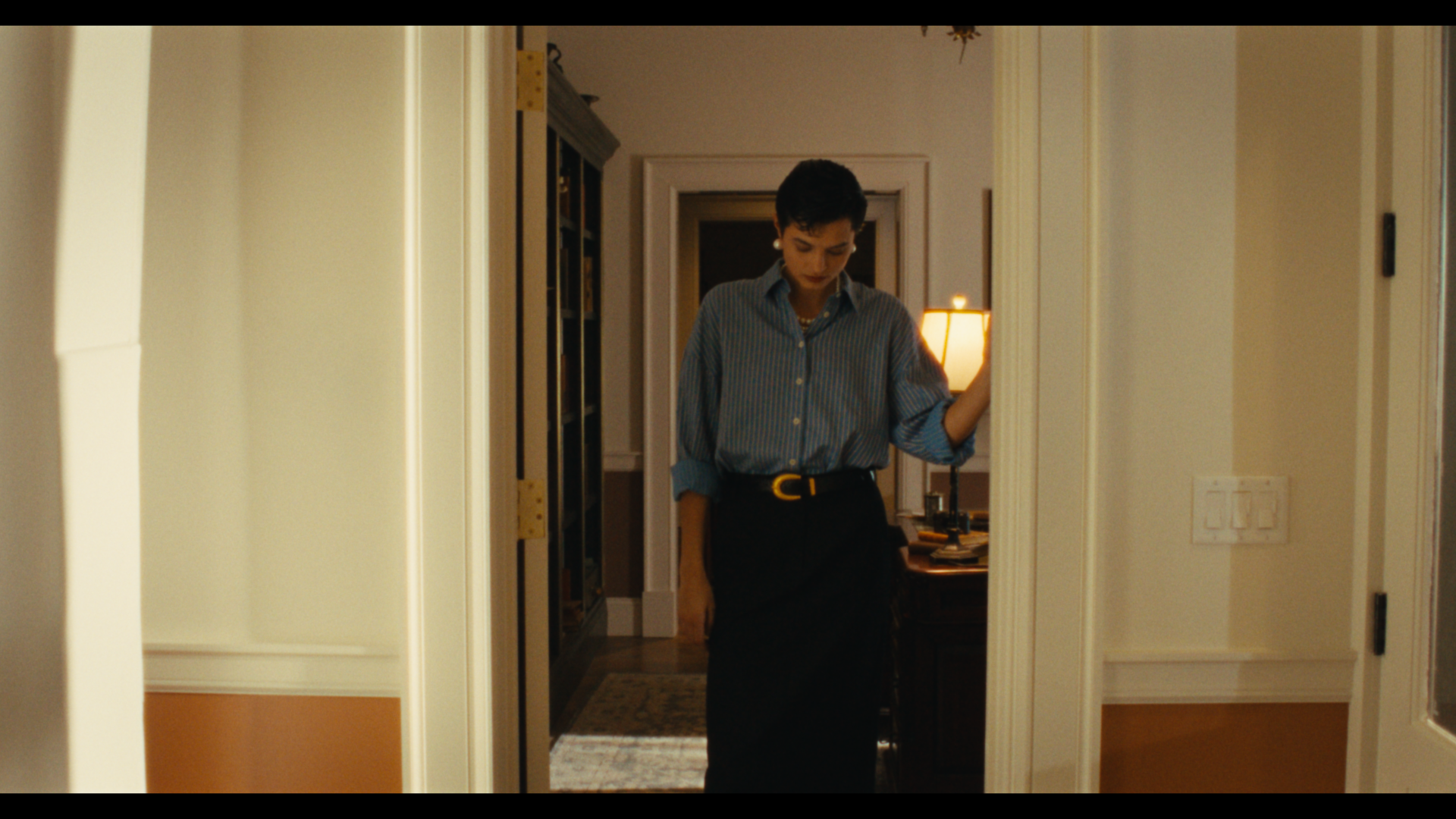 A woman with short dark hair wearing a blue striped shirt and a black skirt stands in a doorway with her head slightly bowed. The background shows a dimly lit room with a table, papers, and a lamp.