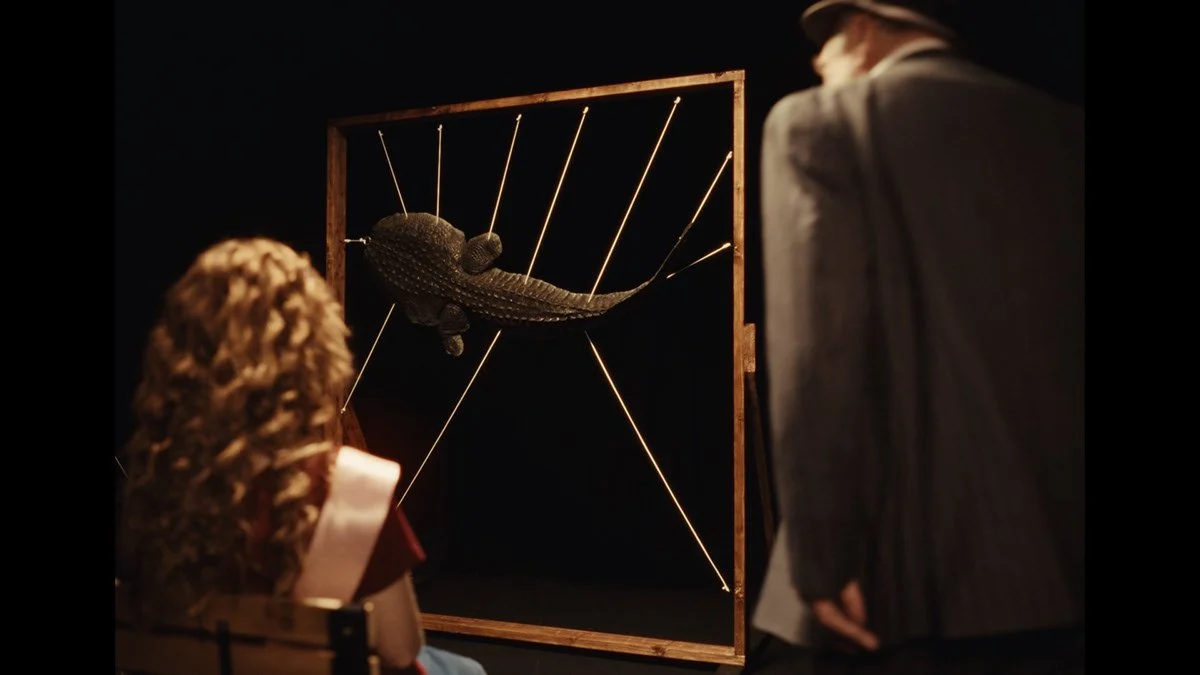 A museum exhibit featuring a crocodile skeleton suspended within a wooden frame with lines radiating outward, while two visitors observe it in a darkened room.