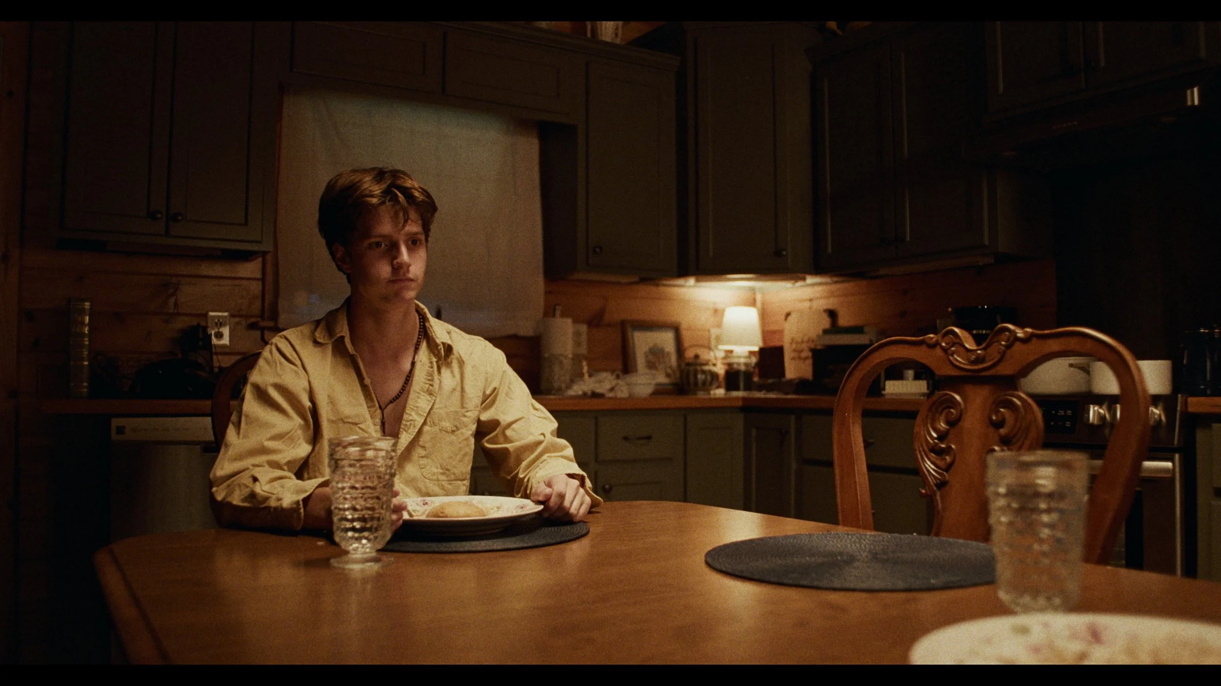 Young man sitting alone at a wooden dining table in a rustic kitchen, with a plate of food and a glass of water, dimly lit by a small lamp. Mathias White: writer, director, cinematographer / director of photography, and editor.
