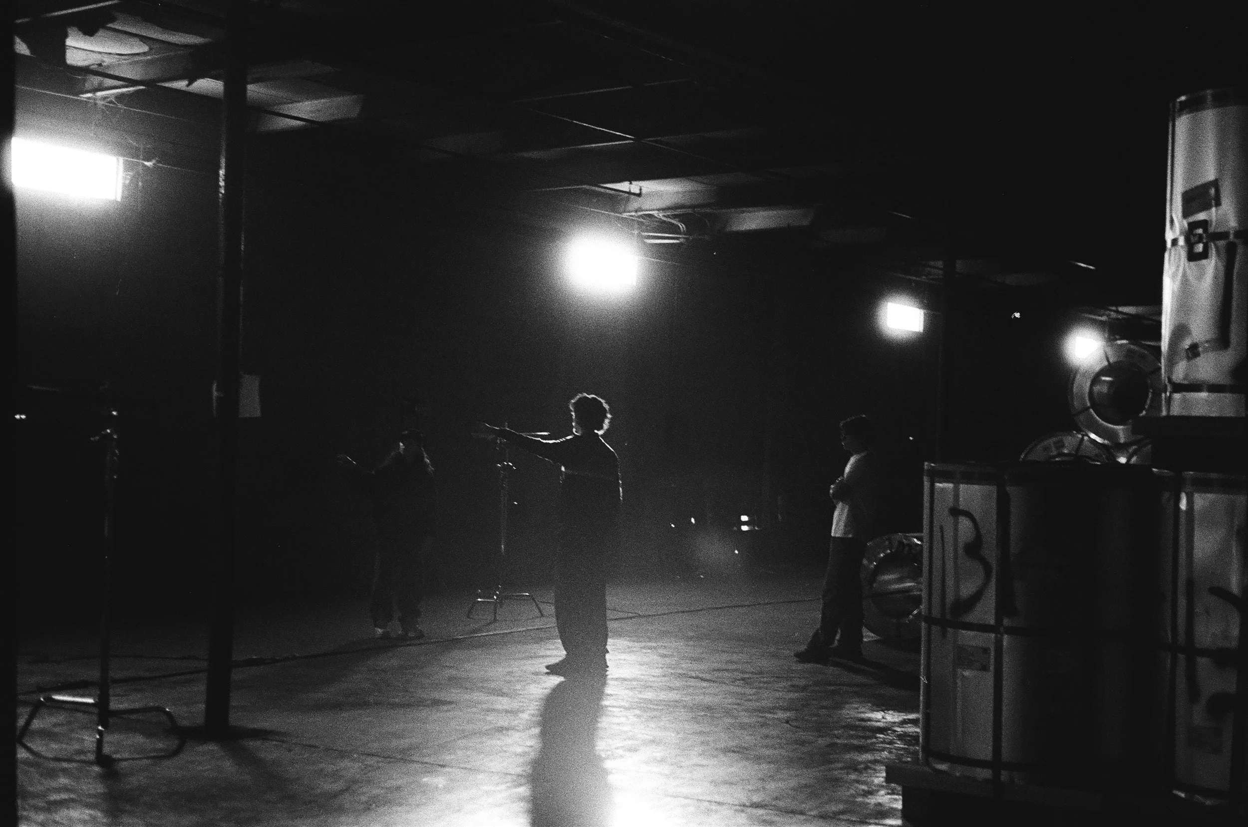 A black and white photo of a dimly lit indoor space with four people standing around. One person appears to be giving directions or a speech, with bright lights above. Industrial objects or equipment are visible, with some containers numbered 3 and 4