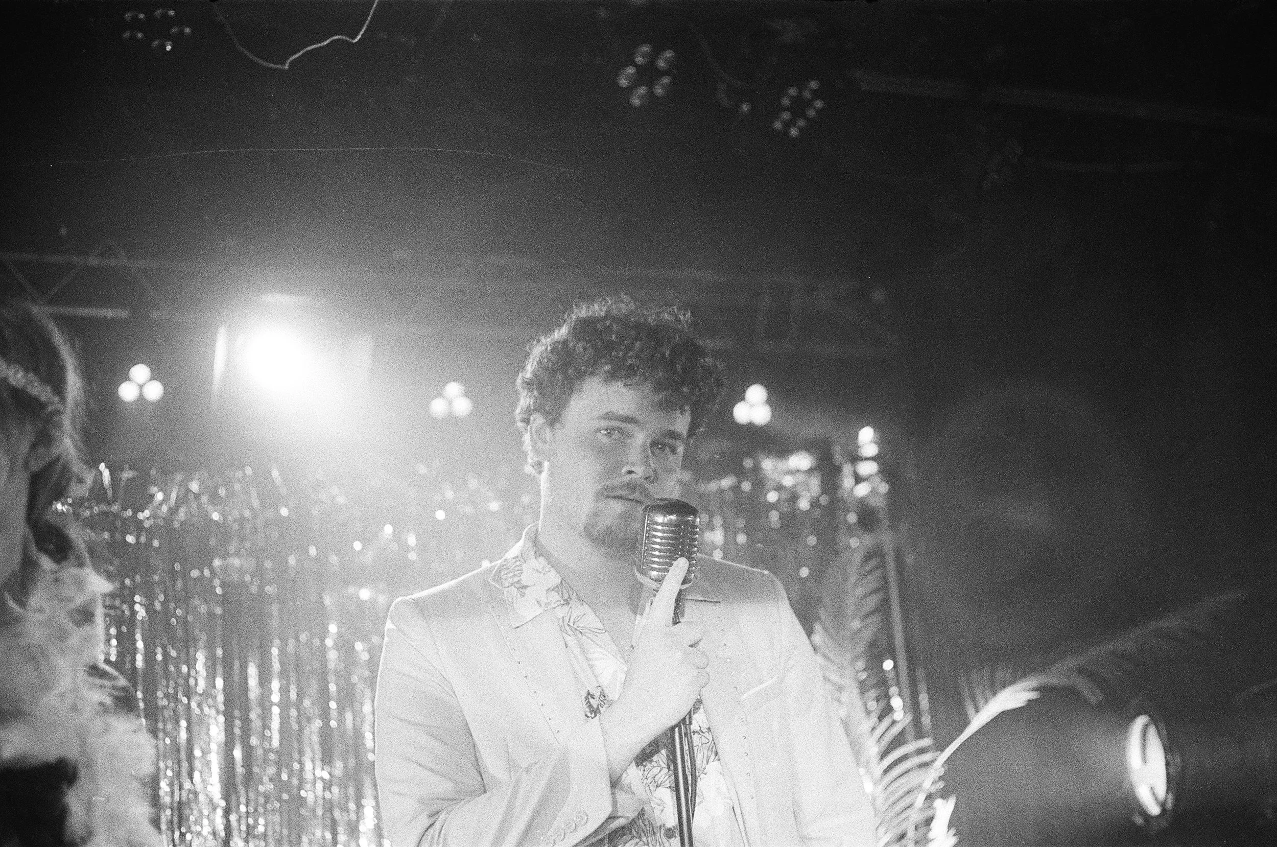 A man with curly hair and a beard, wearing a light-colored blazer and a patterned shirt, stands on stage holding a vintage microphone. Stage lights and shiny decorations are visible in the background.