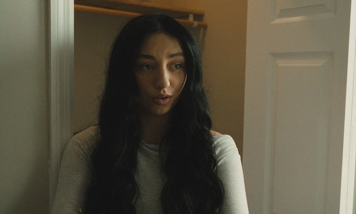 A woman with long black wavy hair and fair skin standing in a room, with her arms crossed, wearing a light-colored top, looking slightly expressions of surprise or concern.