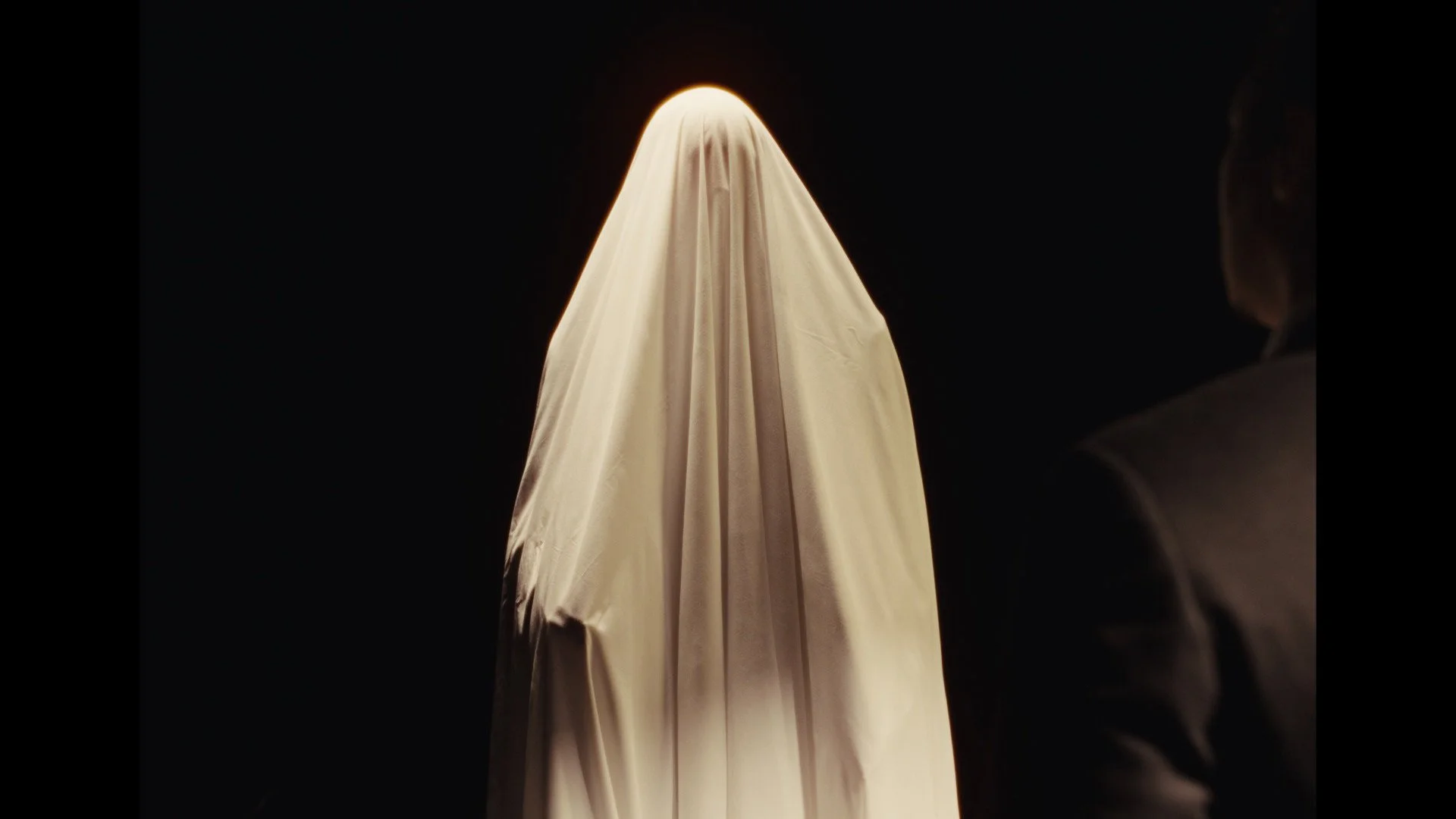 Person in a dark suit watching a figure covered by a white sheet in a black room