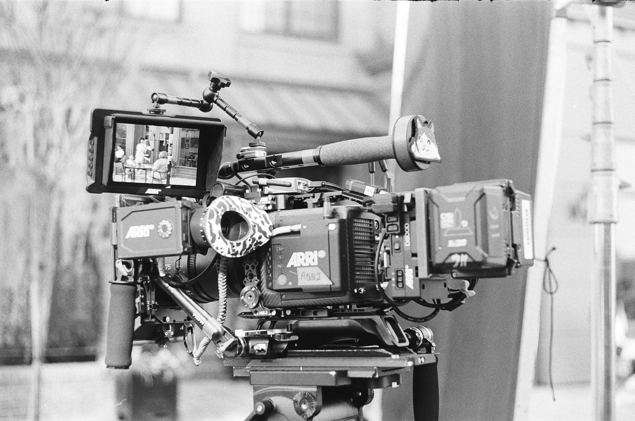 Black and white image of a professional video camera mounted on a tripod, with a small monitor attached showing a scene being filmed.