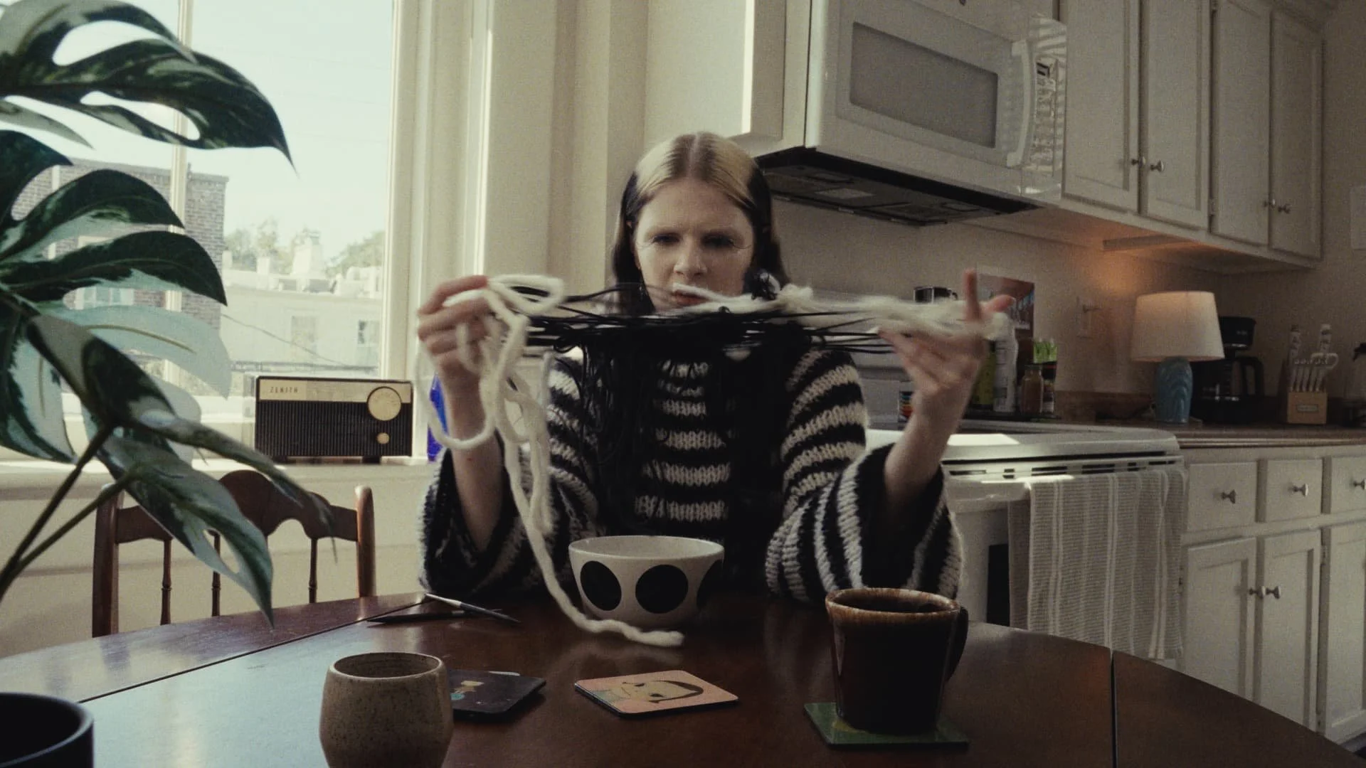 A woman with long dark hair pulled back, wearing a black and white striped sweater, sits at a wooden dining table in a kitchen. She holds a bunch of yarn in her hands and looks perplexed. There are various cups and small items on the table, and a lar