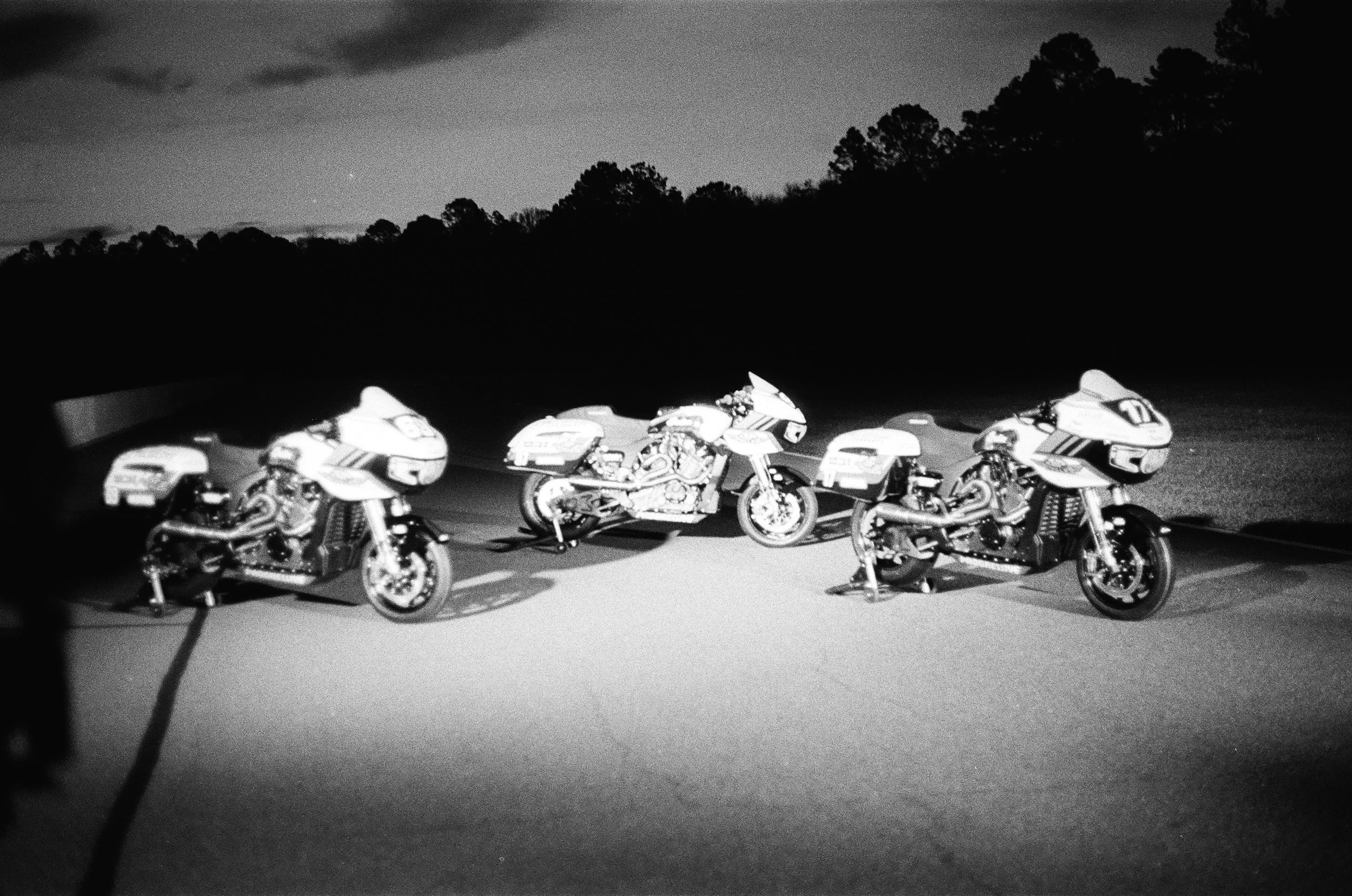 Three motorcycles parked on a road at dusk with trees in the background, black and white photo.