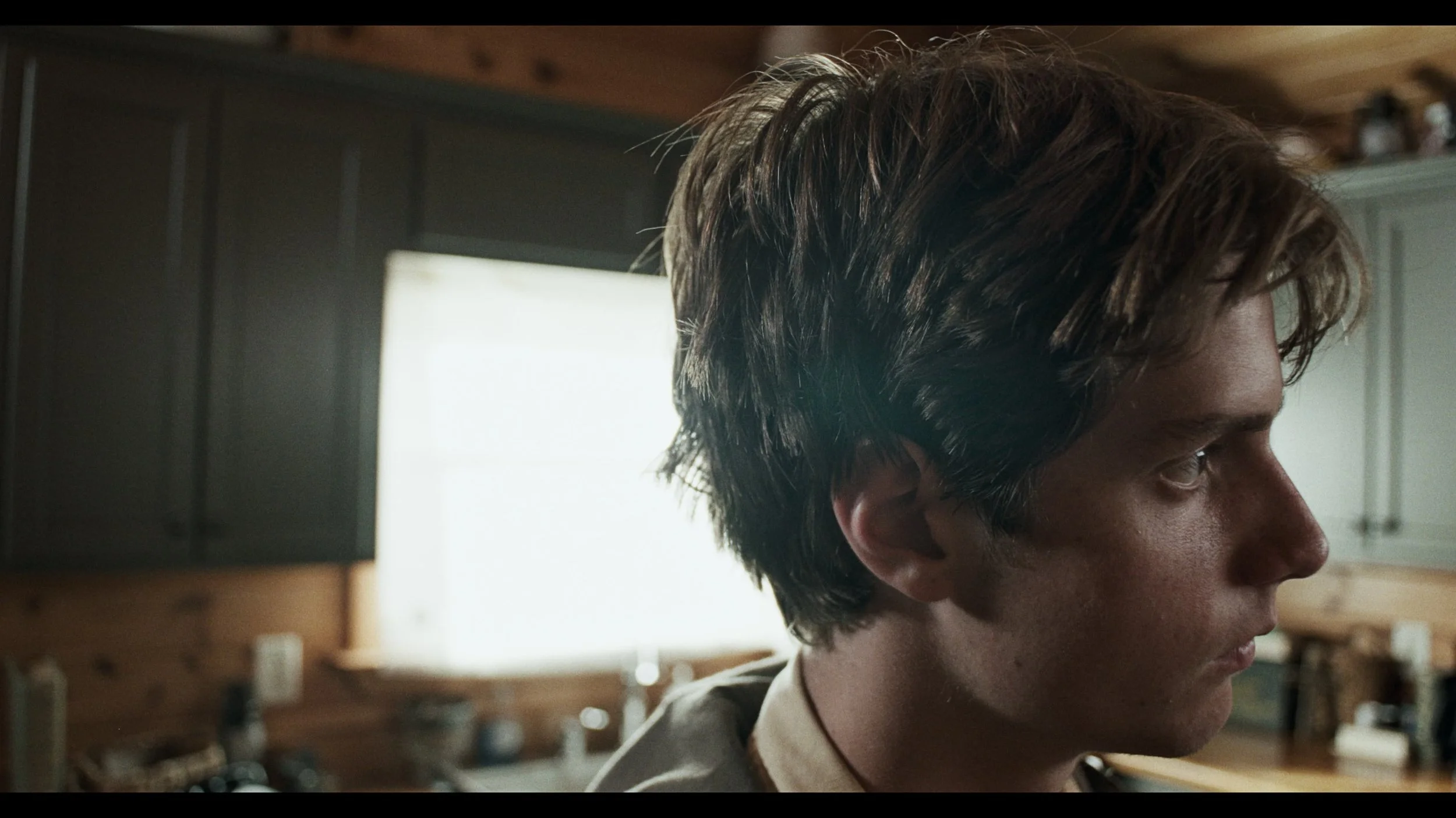 A man with wavy brown hair looking to the right in a room with wood paneling and a window in the background.