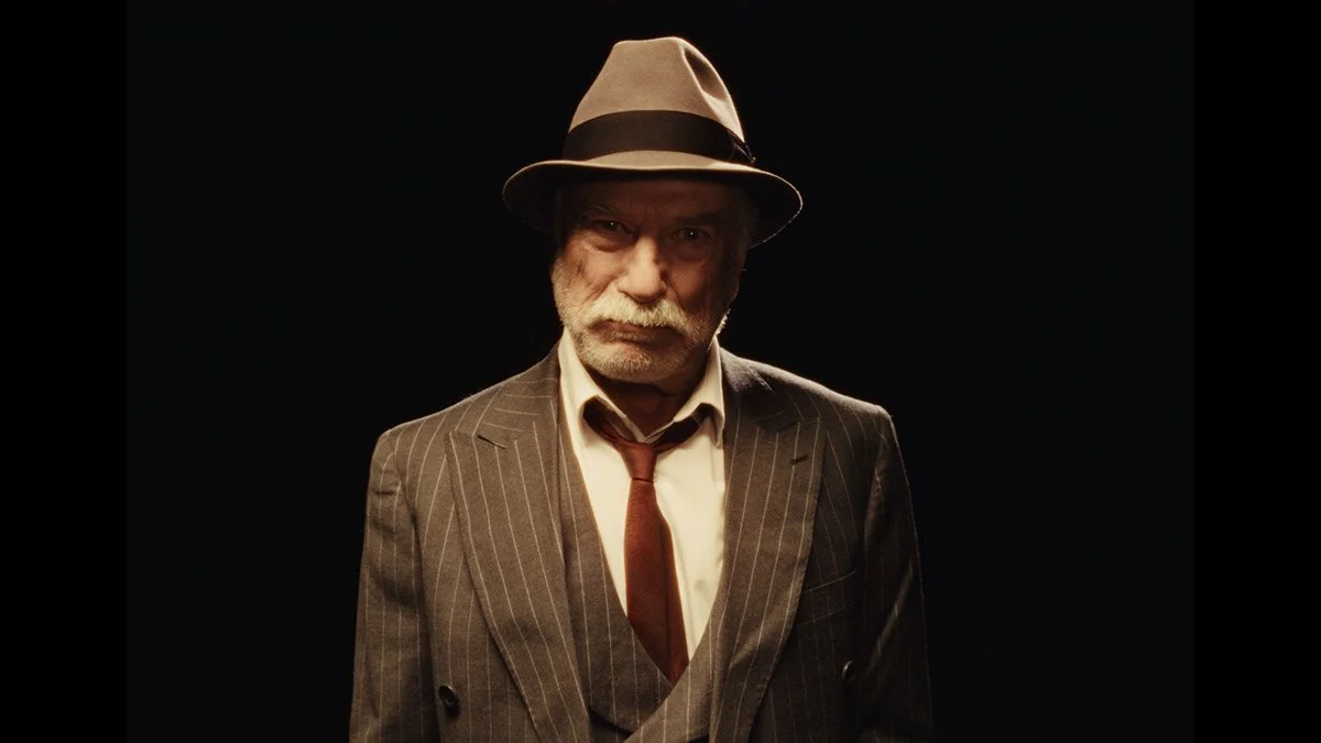 An older man with a white beard and mustache wearing a fedora hat, a pinstripe suit, a white shirt, and a red tie, standing against a black background.