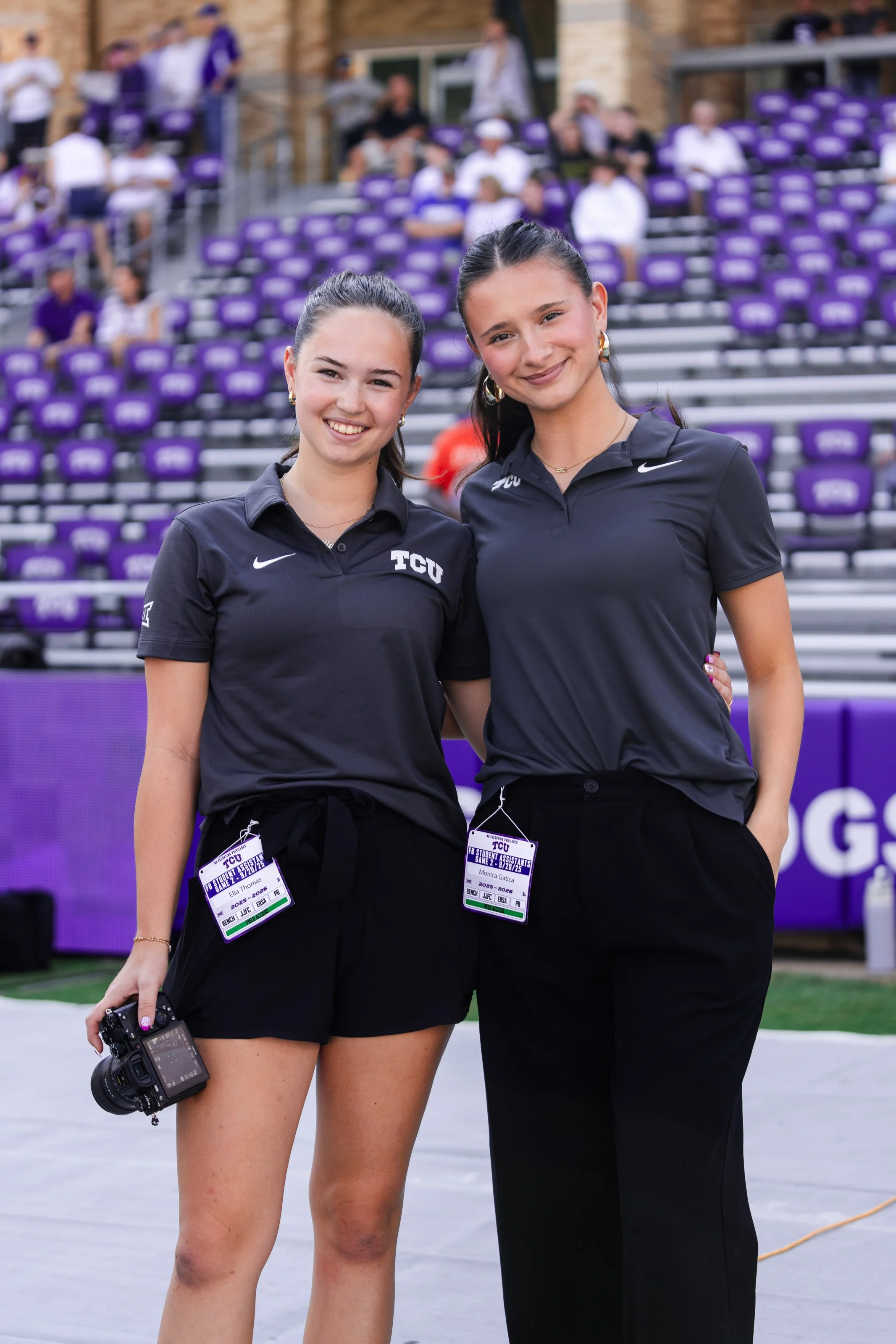 Two young women with dark hair, wearing black TCU polo shirts and black shorts or pants, standing outdoors on a sports field or court, smiling at the camera, with purple stadium seating and spectators in the background.