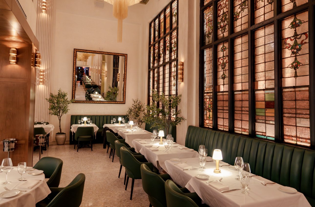 A beautifully decorated restaurant dining area with white tablecloths, green chairs, a large mirror on the wall, potted plants, and stained glass windows.